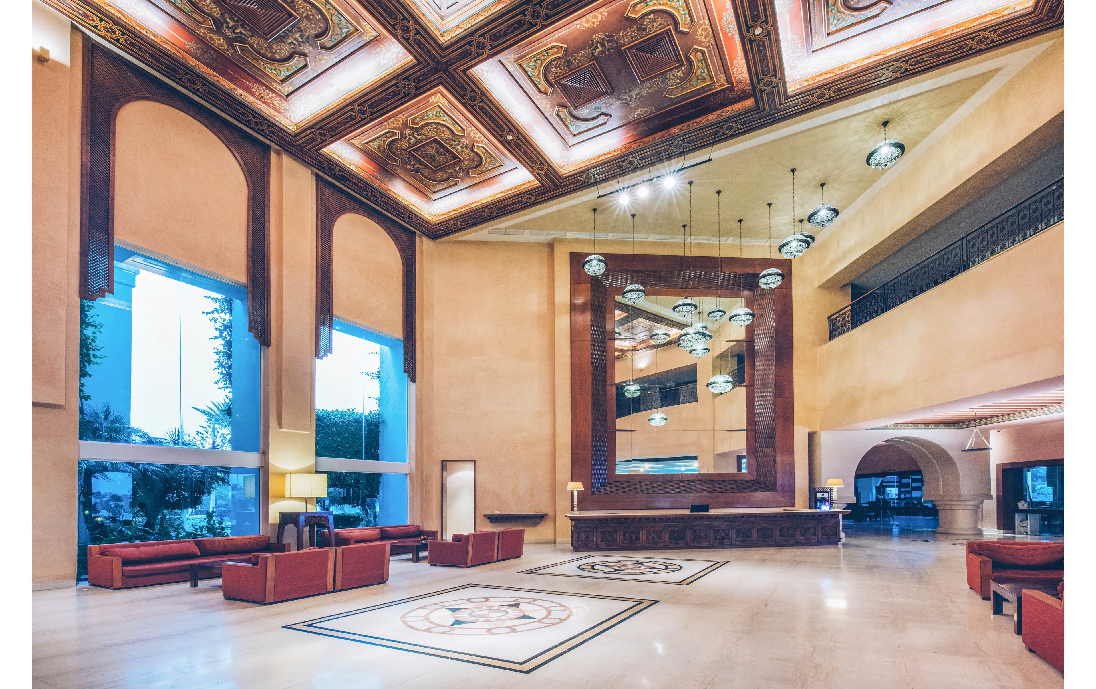 A large, elegant hotel lobby features high ceilings and warm lighting. Ornate wooden panels with intricate designs decorate the ceiling above the reception area. Modern pendant lights hang in front of a tall mirrored wall behind the front desk. Red upholstered sofas and armchairs are arranged across the polished floor, with large windows bringing in natural light from outside.