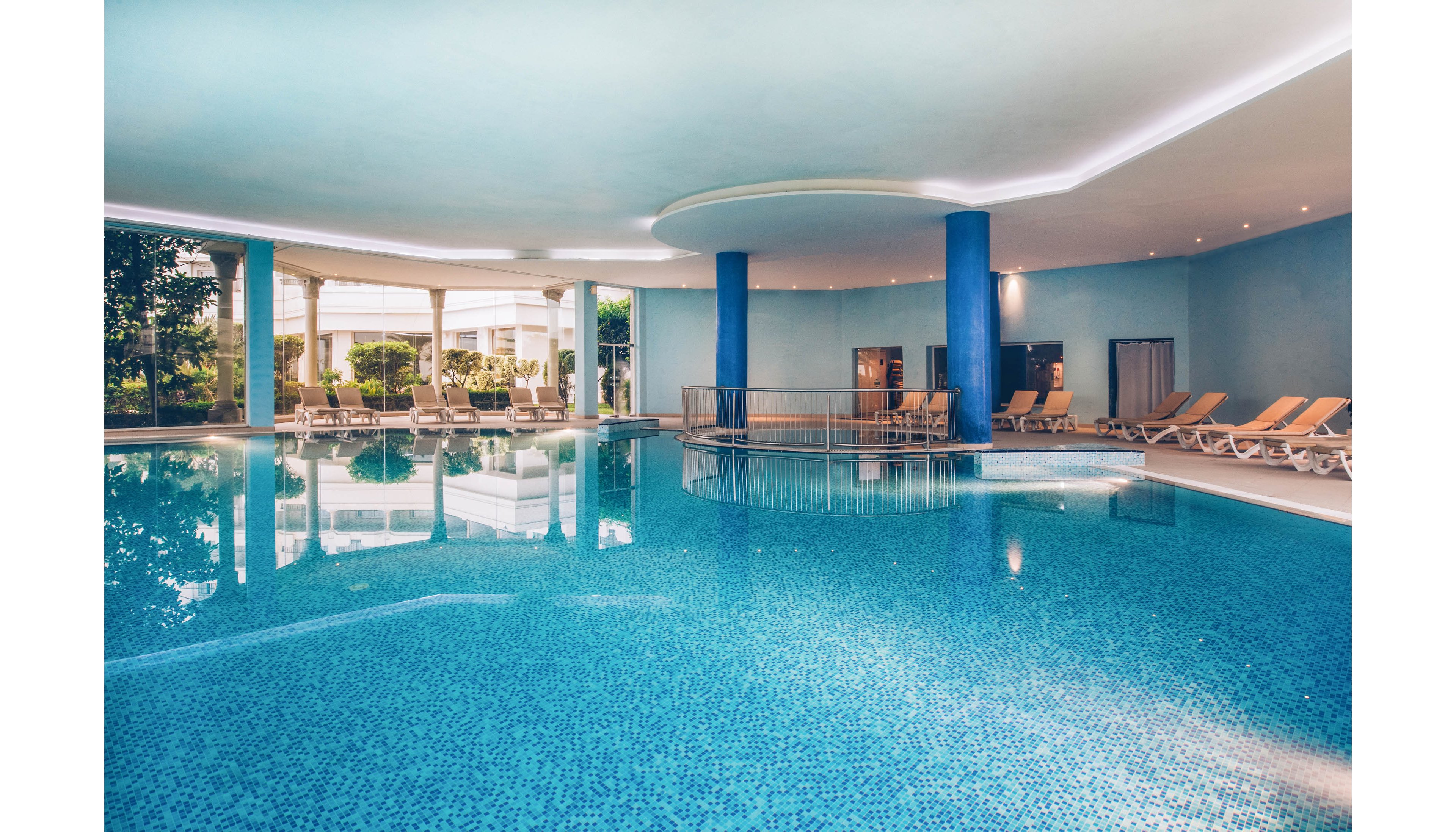 Spacious indoor swimming pool with clear blue water in a contemporary hotel or spa setting. Beige lounge chairs line the poolside, offering a relaxing area for guests. Large blue columns and soft ceiling lighting create a calm, modern atmosphere. Floor-to-ceiling windows open to a green outdoor area, adding natural light and a resort-like feel.