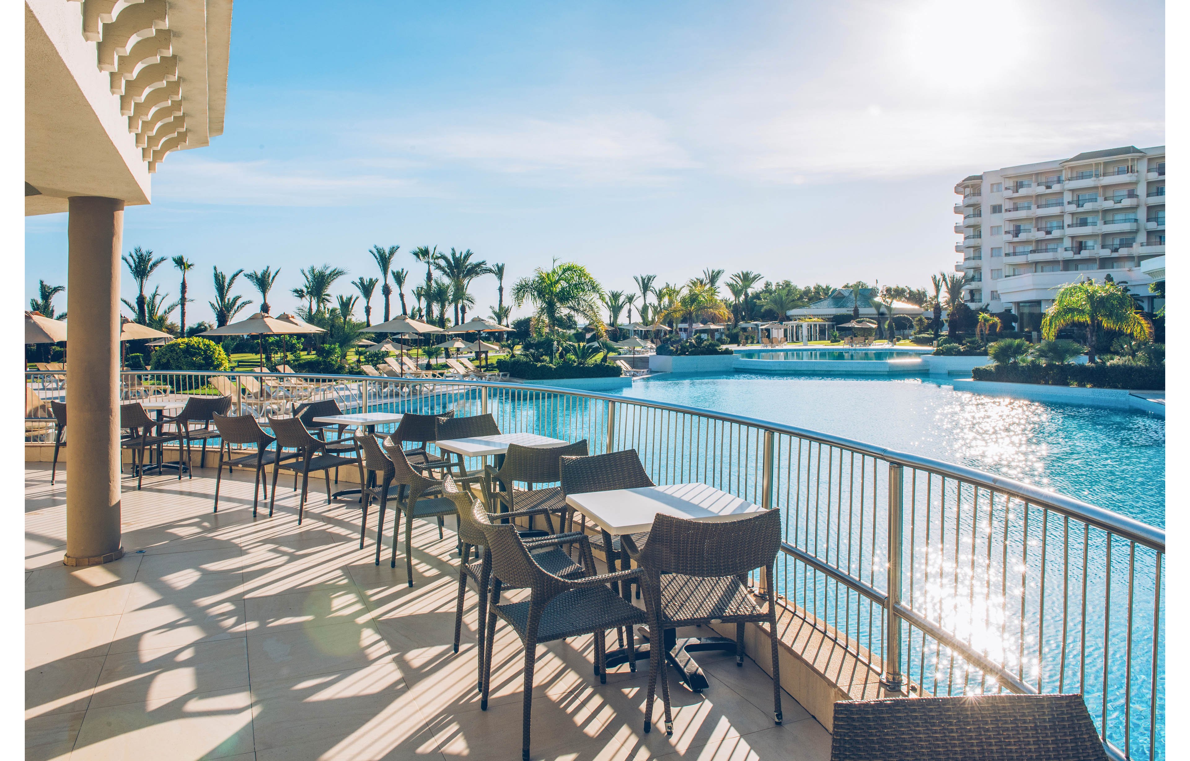 An outdoor terrace cafe overlooks a large resort swimming pool on a sunny day. Empty tables and wicker chairs line the railing, casting long shadows across the tiled floor. Palm trees, sun loungers, and umbrellas surround the expansive pool, with modern hotel buildings in the background. The bright blue water and clear sky create a relaxed, vacation atmosphere.