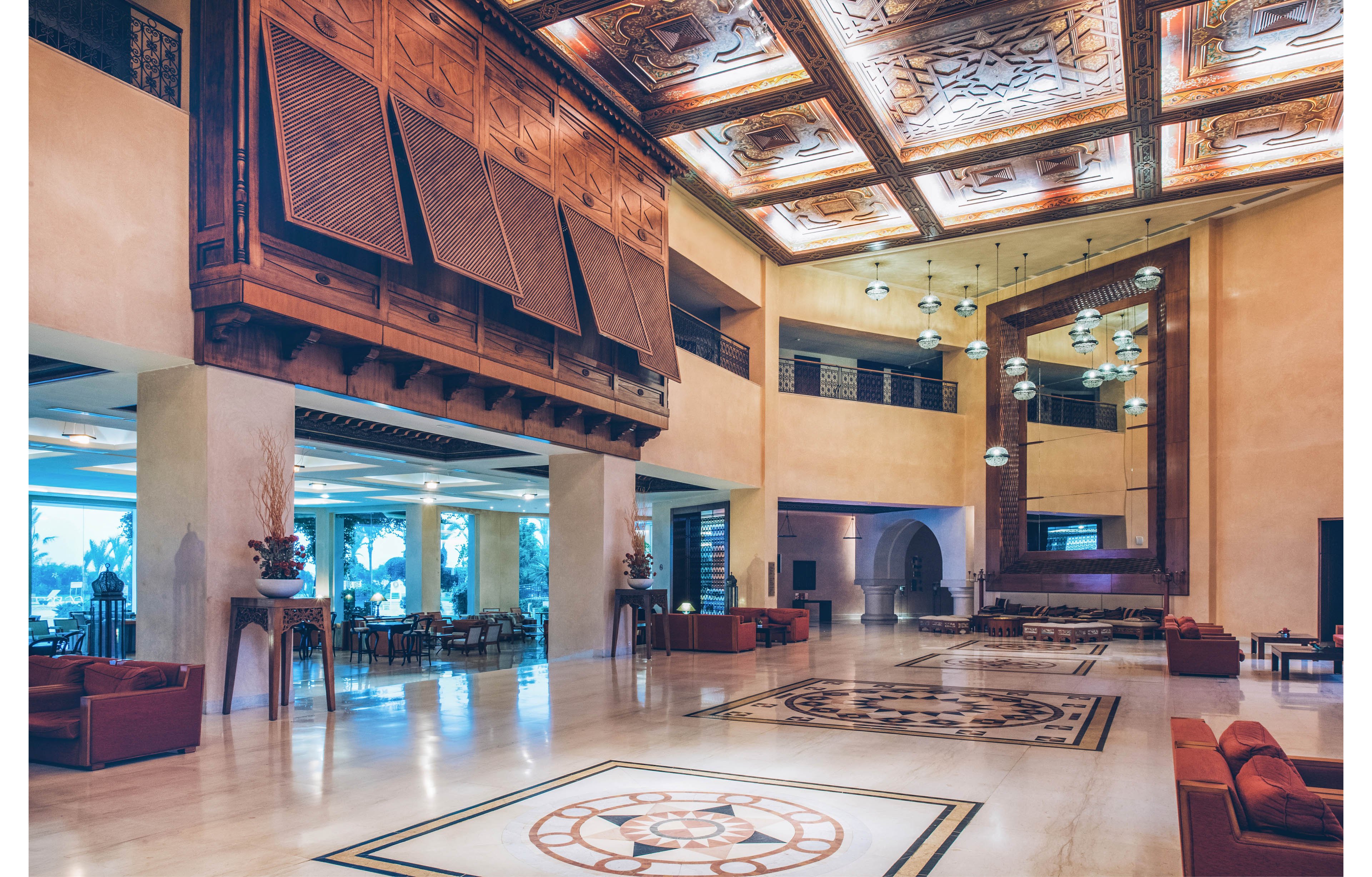 Spacious hotel lobby featuring an ornate carved wooden ceiling and large decorative panels. The polished marble floor showcases intricate geometric inlays that echo the ceiling design. Warm ambient lighting and hanging pendant lamps create an inviting atmosphere. Comfortable leather seating areas and tall vases with dried arrangements complete the elegant interior setting.