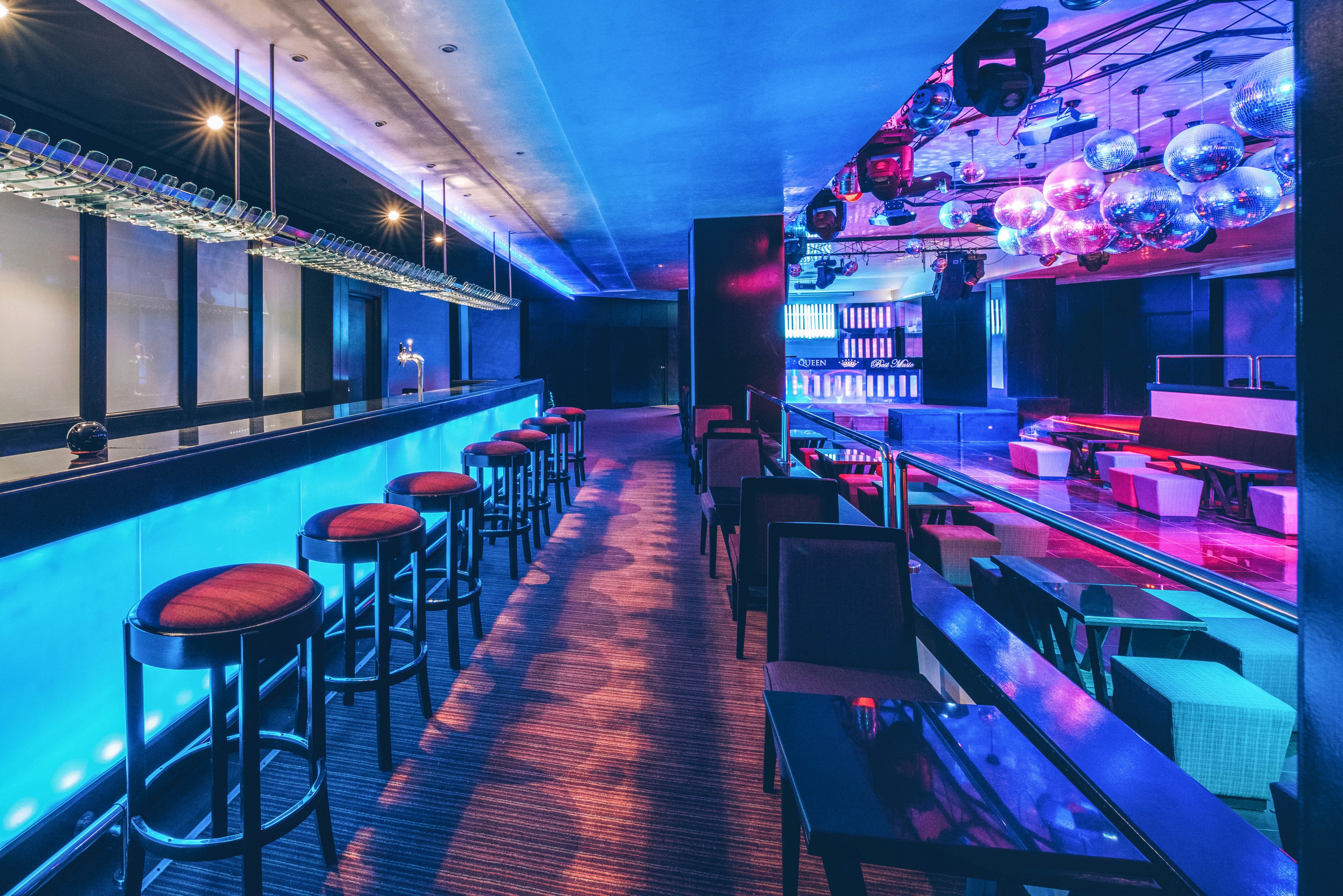 A sleek, contemporary nightclub interior features a long illuminated bar lined with high stools. The space is empty, showcasing colorful blue and pink neon lighting that reflects off glossy surfaces. On the right, a dance floor and lounge seating area extend toward a DJ booth and shelves of backlit bottles. Disco balls and stage lighting fixtures hang from the ceiling, enhancing the vibrant nightlife atmosphere.