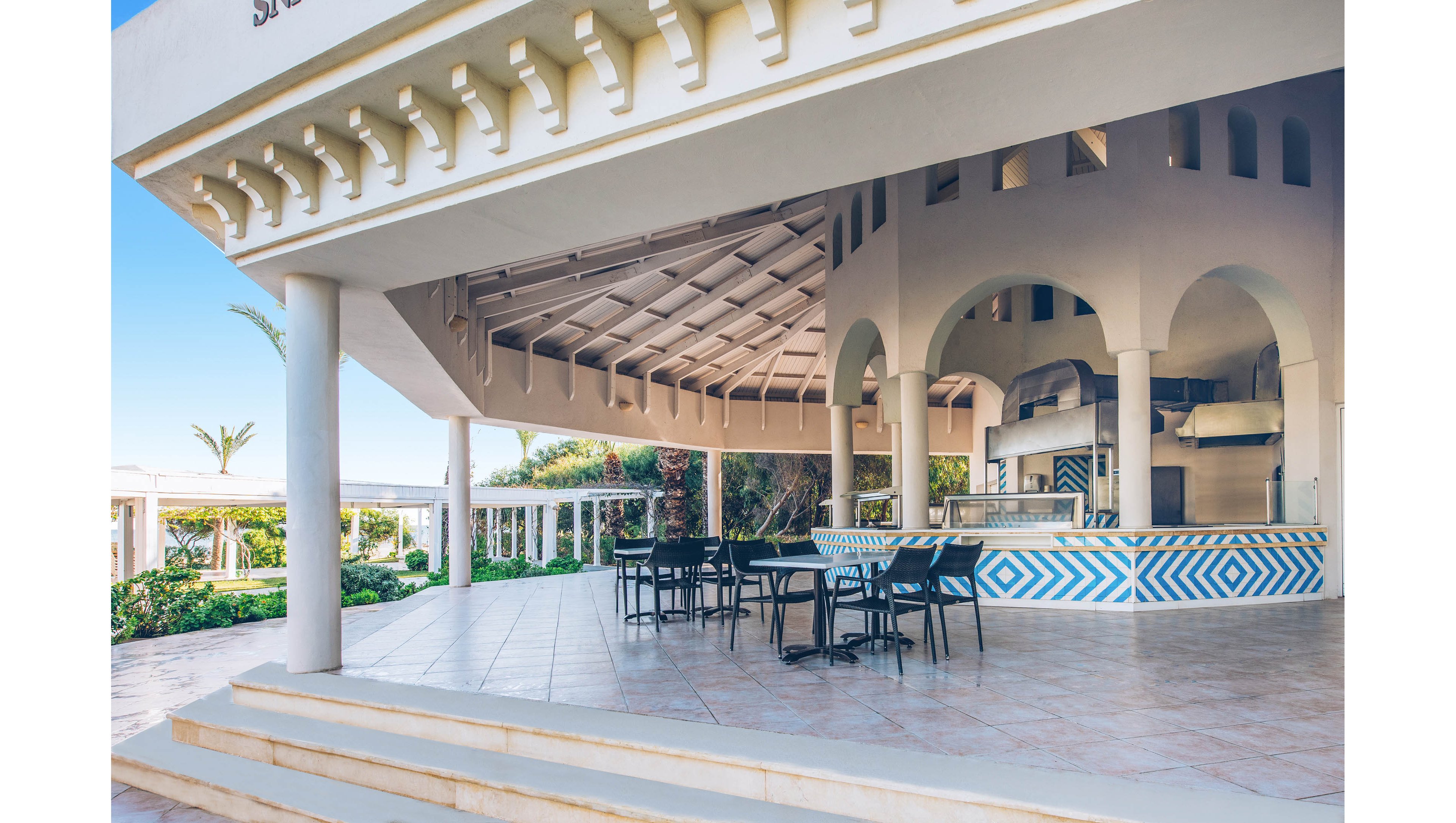 An open-air bar area at a resort features a circular counter with a blue and white chevron pattern. Several empty tables and chairs are arranged on a tiled terrace under a large, curved roof. Arched openings reveal the bar equipment and a glimpse of the surrounding greenery. The scene is brightly lit with clear blue sky, suggesting a warm, relaxed vacation atmosphere.