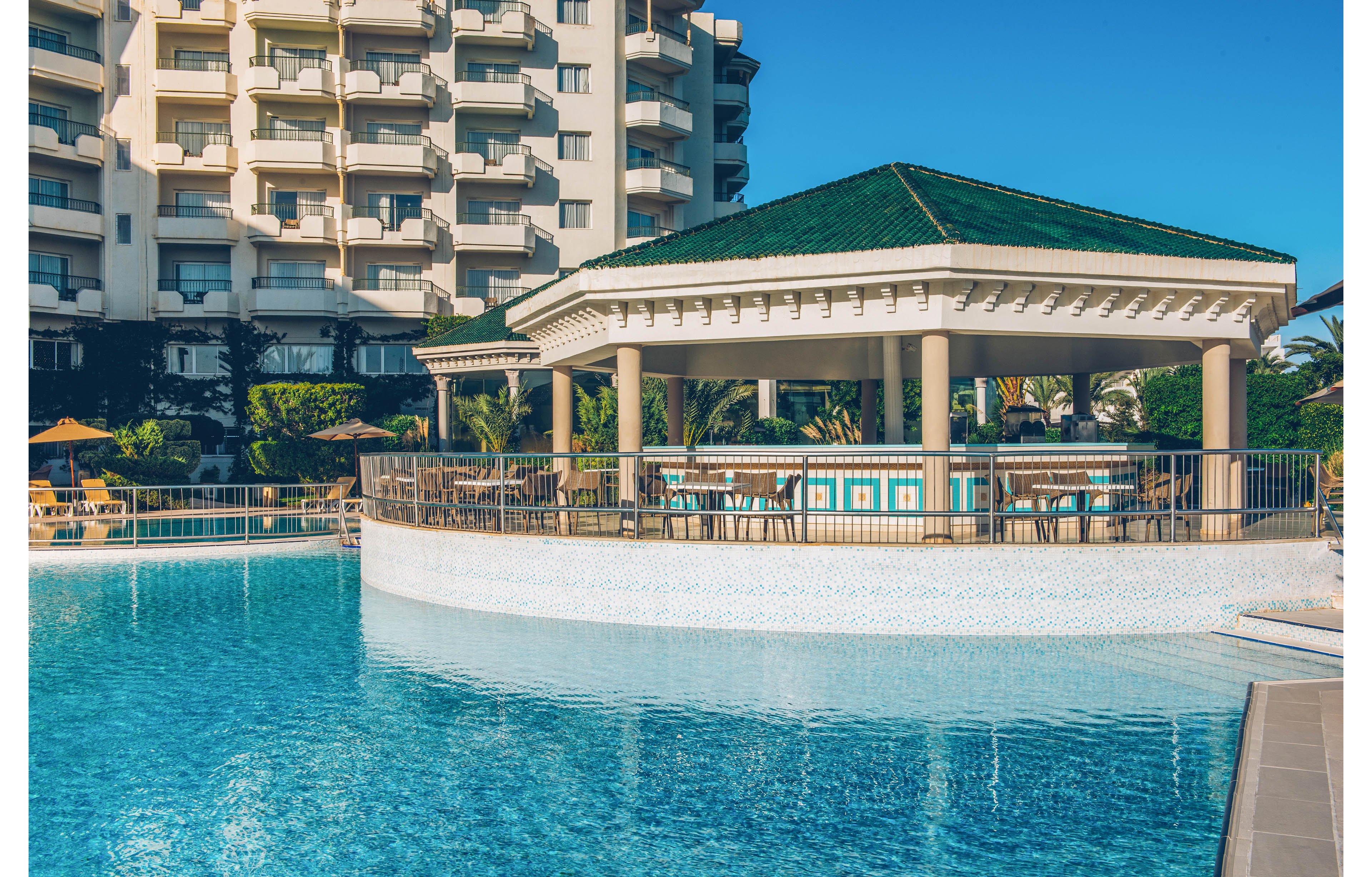 A large resort swimming pool surrounds an open-air bar pavilion with a green tiled roof. The structure is supported by columns and enclosed by railings, sitting directly over the water. In the background, a multi-story hotel building with balconies rises against a clear blue sky. Palm trees and sun loungers suggest a warm, relaxing vacation setting. The calm water reflects the architecture and bright daylight.