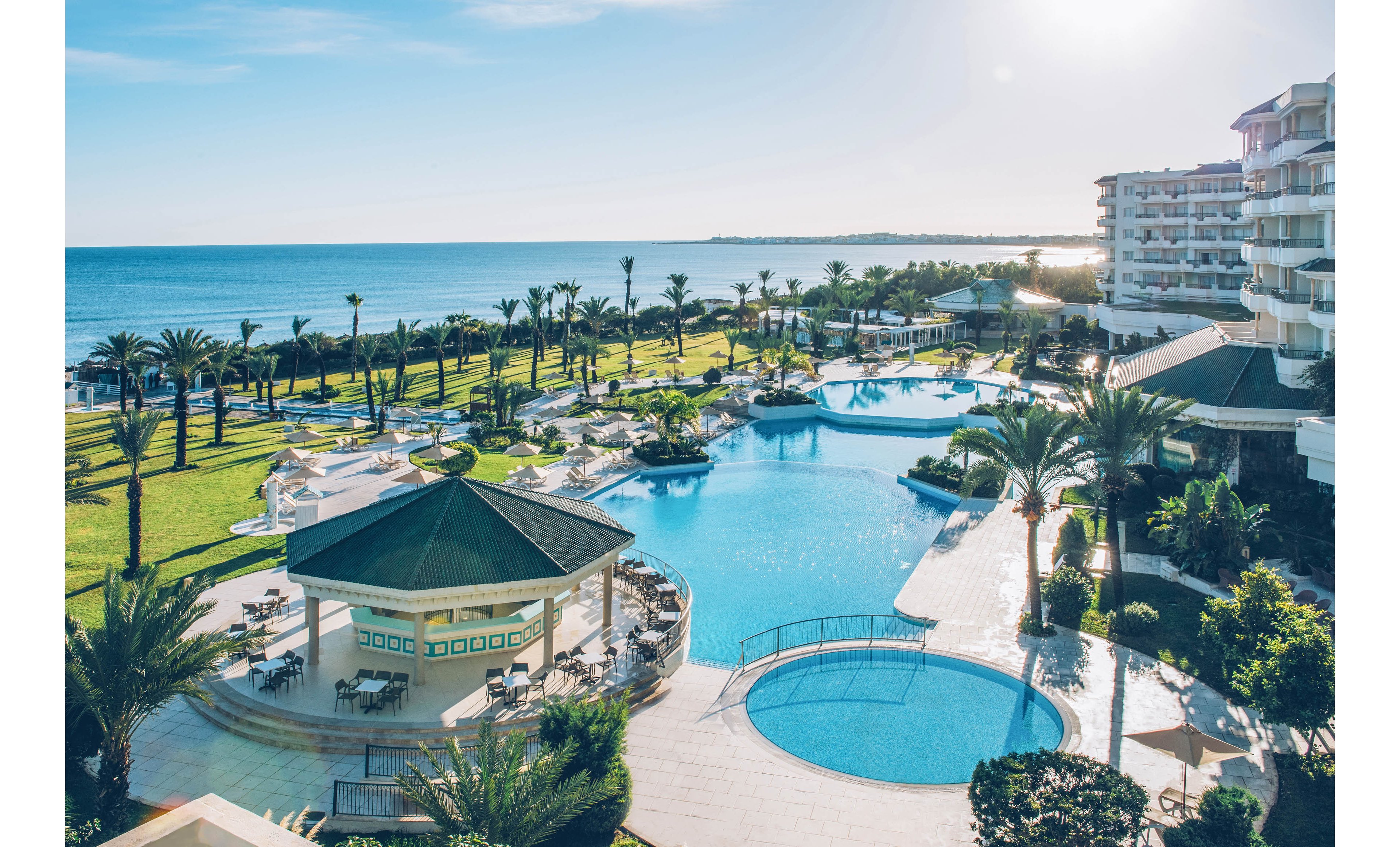 A large coastal resort features an expansive swimming pool complex overlooking the sea. Multiple pools, sun loungers, and shaded cabanas are arranged among palm trees and manicured lawns. A central pool bar with a green roof sits beside curved walkways and terraces. Modern multi-story hotel buildings frame the scene under a clear blue sky, creating a relaxed vacation atmosphere.
