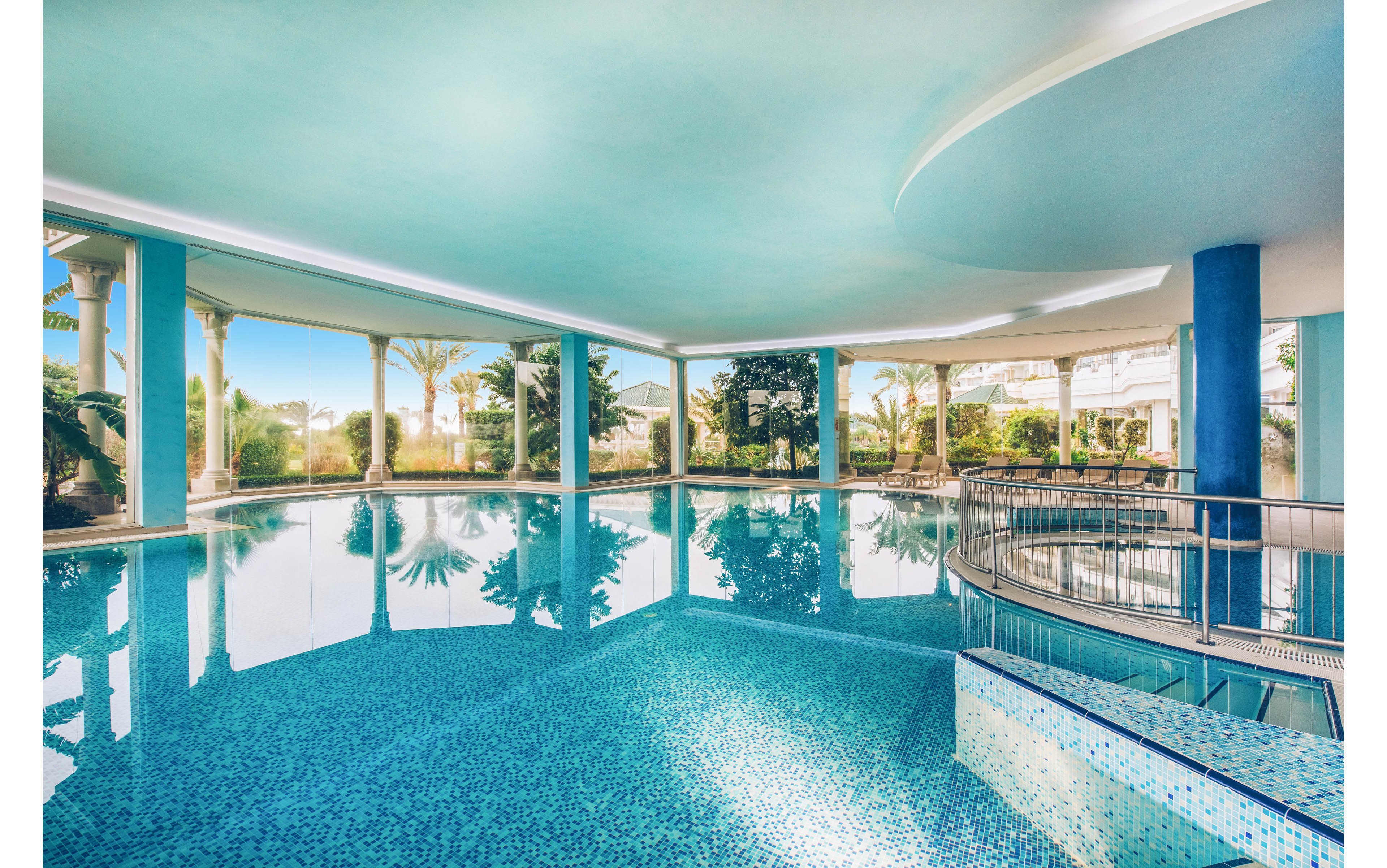 A spacious indoor swimming pool features shimmering blue mosaic tiles and calm, reflective water. Large open arches reveal a lush outdoor garden with palm trees and bright daylight. Elegant white columns and a curved railing add a sophisticated architectural touch. The serene atmosphere suggests a high-end spa or resort setting ideal for relaxation and leisure.