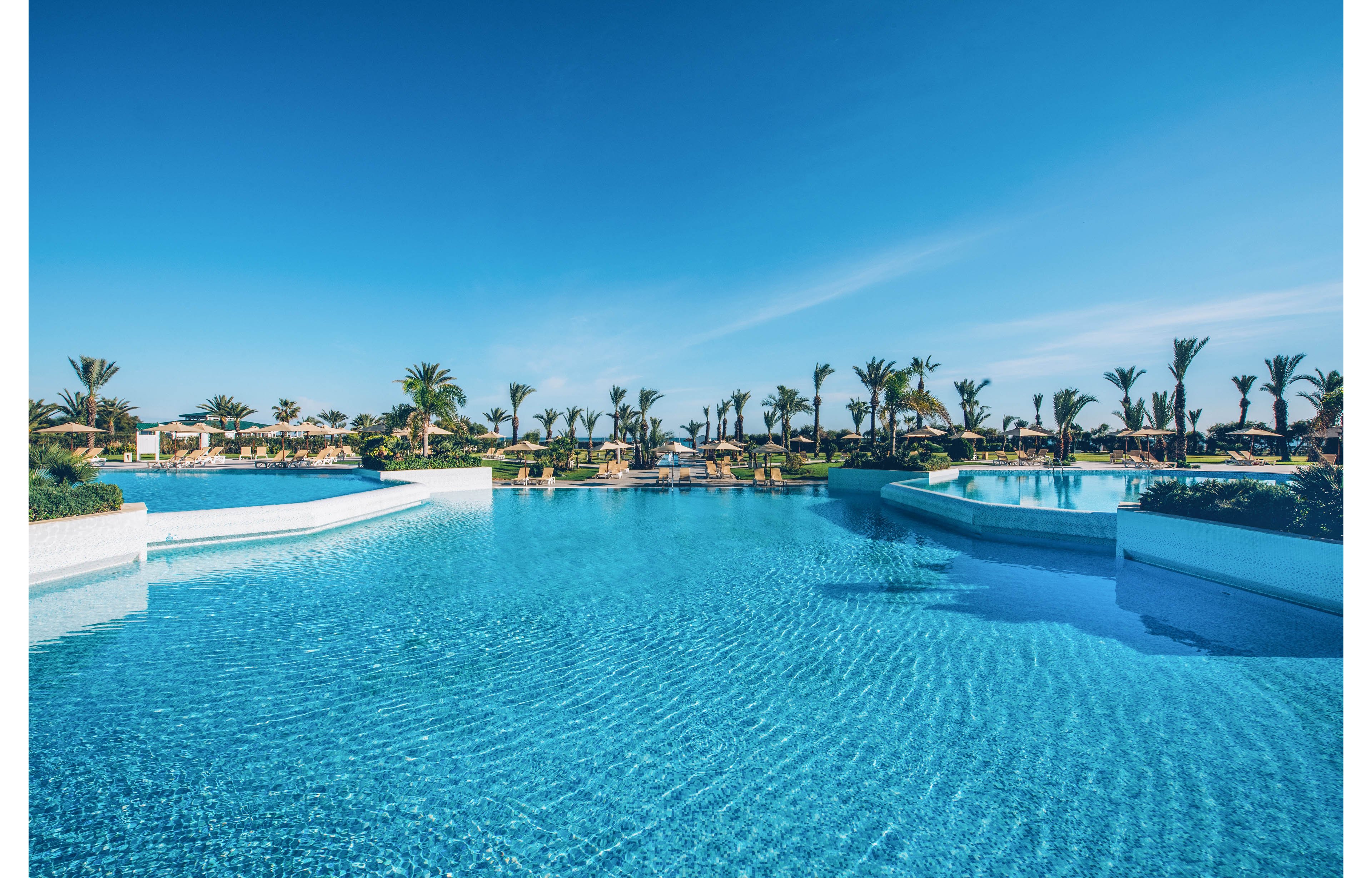 A large, crystal-clear swimming pool stretches across the foreground under a bright blue sky. Palm trees and sun loungers line the distant edge of the pool, suggesting a tropical resort setting. The calm water surface reflects the sky, creating a serene and inviting atmosphere. The overall scene conveys relaxation, vacation, and warm weather leisure.