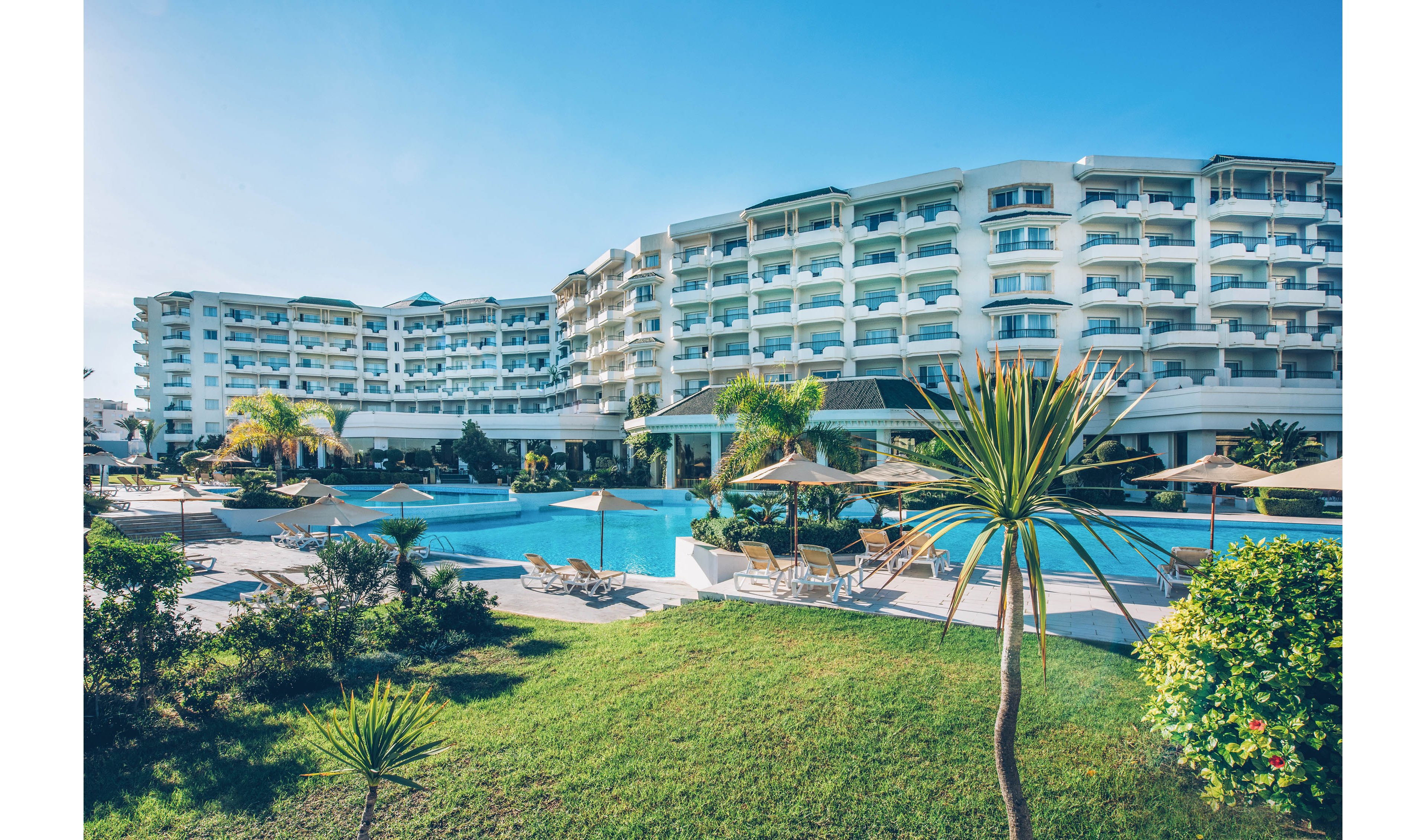 A large modern resort complex with multiple white buildings surrounds a central outdoor swimming pool. Sun loungers and parasols are arranged neatly around the pool, suggesting a relaxing vacation atmosphere. Lush green lawns, palm trees, and tropical plants add to the feeling of a warm coastal destination. The clear blue sky and bright sunlight create a serene and inviting holiday scene.