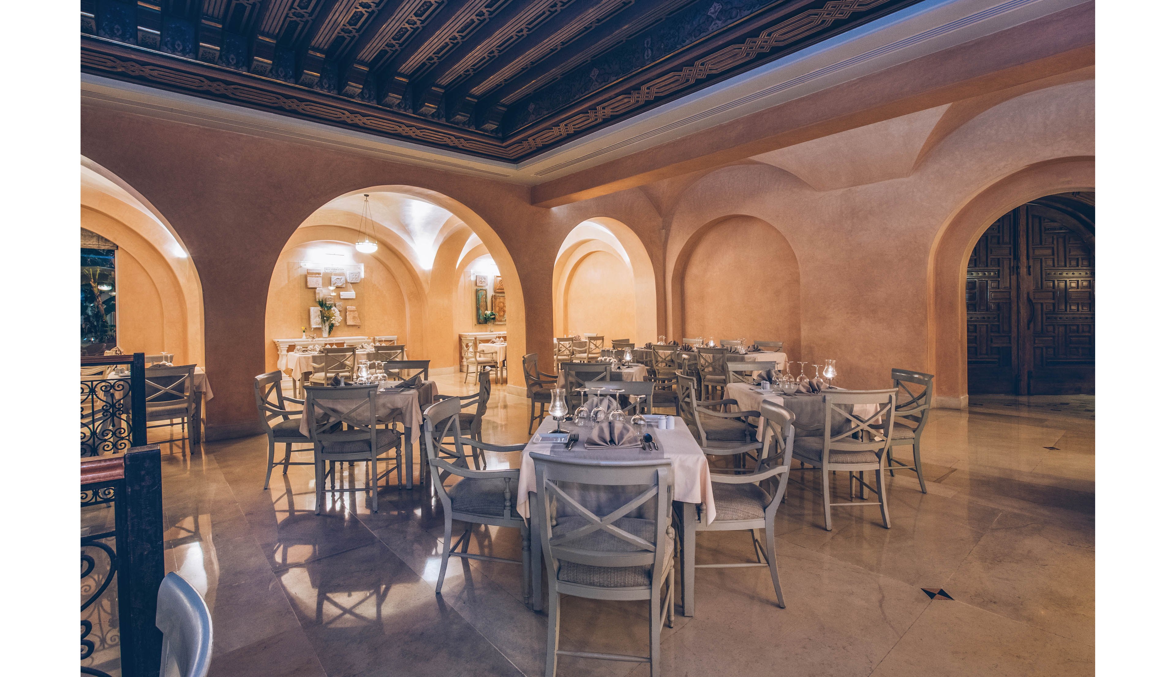 An upscale restaurant dining room is set with neatly arranged tables and chairs, ready for guests. The space features warm beige walls, large arched openings, and a polished stone floor that reflects the ambient lighting. Chandeliers and wall lights create a soft, inviting glow over the white tablecloths and glassware. The overall atmosphere feels calm, refined, and intimate, suggesting a fine dining experience.