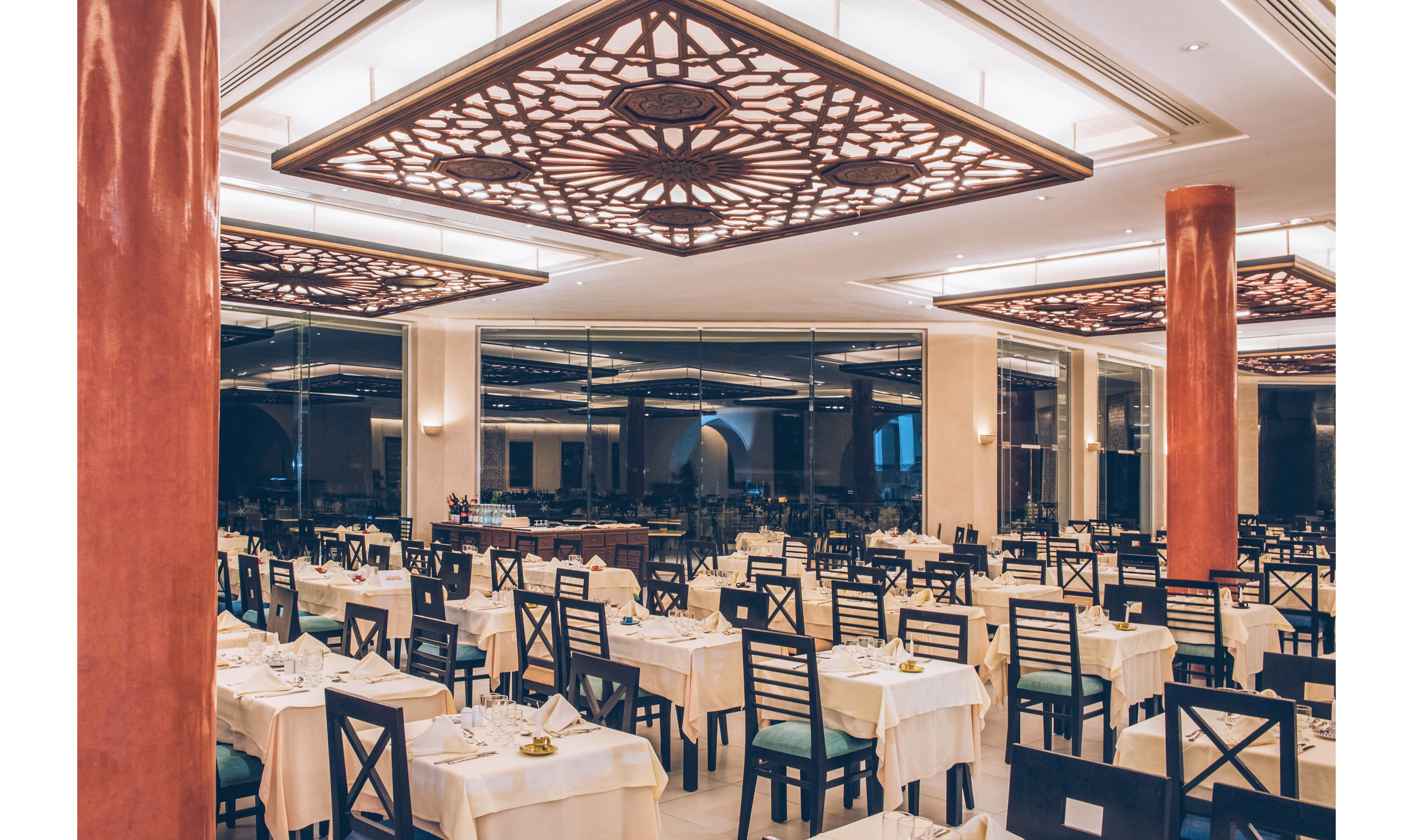 A spacious fine dining restaurant is shown with neatly arranged tables covered in white linens. Dark wooden chairs with cushioned seats are evenly spaced, creating a formal and organized layout. Large decorative ceiling panels with intricate patterns and warm lighting illuminate the room. Floor-to-ceiling windows reflect the interior, adding depth and a sophisticated evening ambiance.