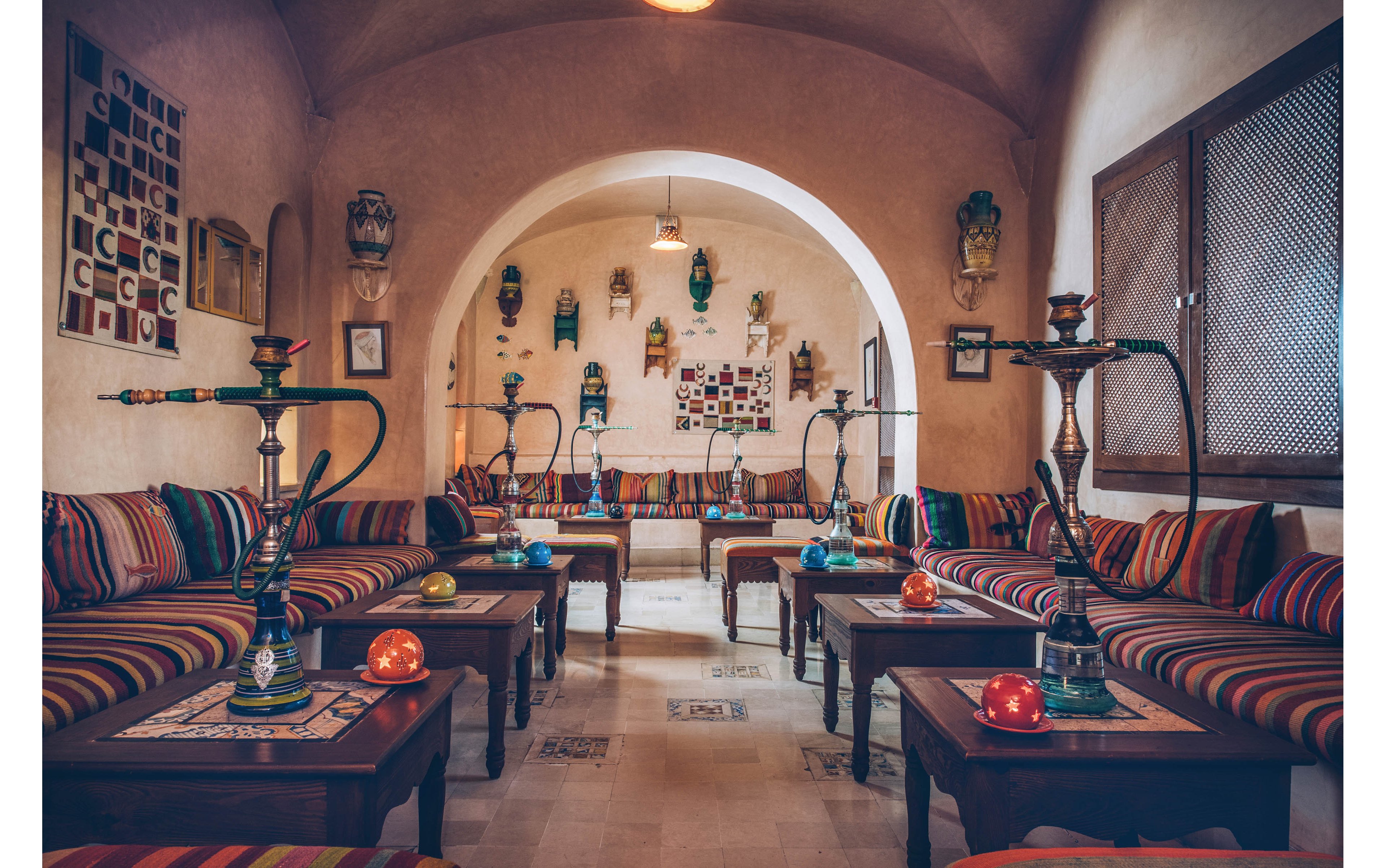 A warm, traditional hookah lounge features multiple shisha pipes arranged on low wooden tables. Colorful striped cushions line the benches along both sides of the room, creating an inviting seating area. Decorative wall art and hanging ornaments accent the arched doorway and cream-colored walls. Soft, ambient lighting enhances the cozy, relaxed atmosphere of the interior space.