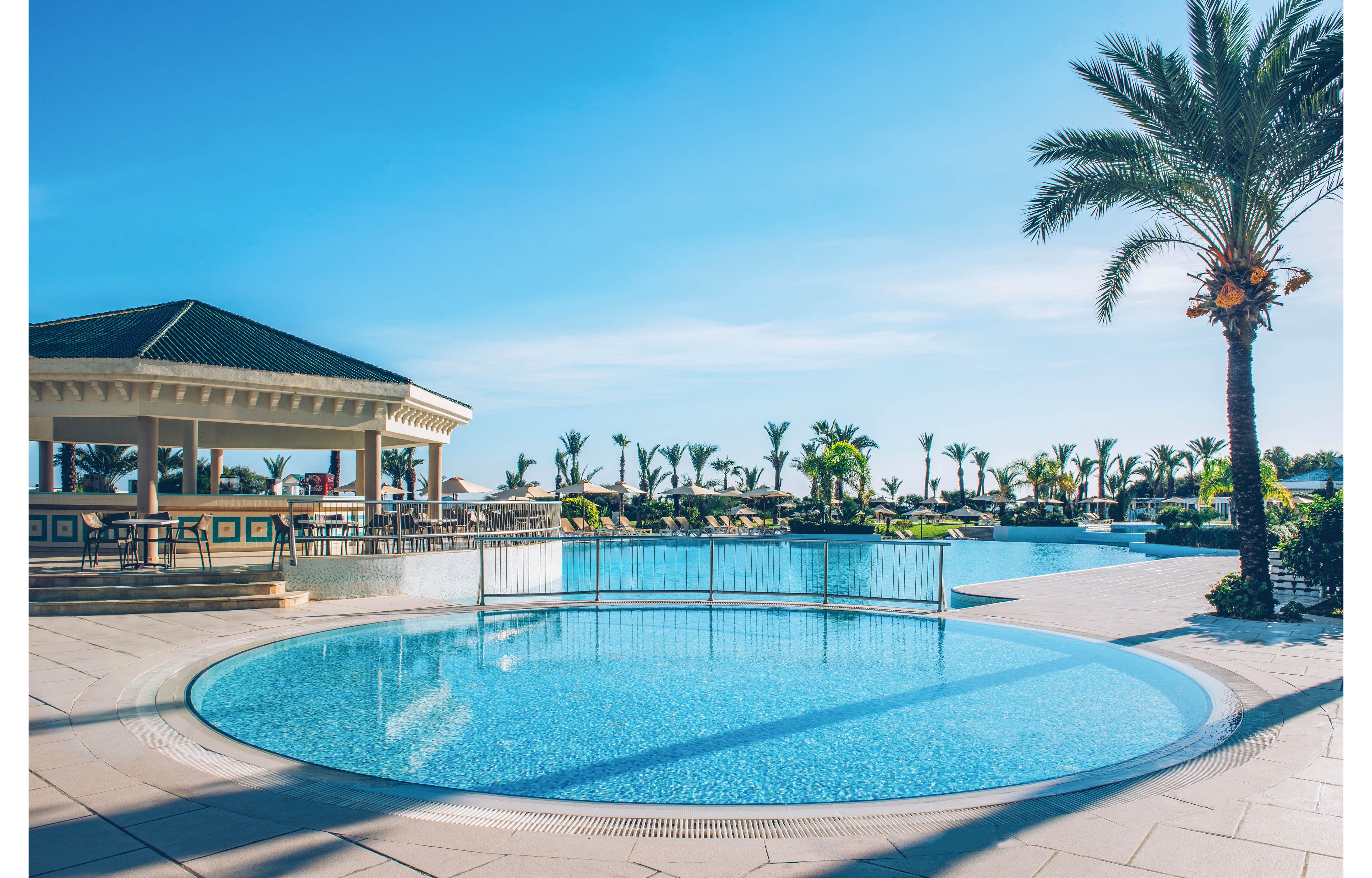 A bright, circular swimming pool sits in the foreground of a tropical resort. Palm trees and sun loungers surround the larger main pool in the background. A covered poolside bar or pavilion stands to the left under a clear blue sky. The scene suggests a warm, relaxing vacation atmosphere with clean, modern architecture.