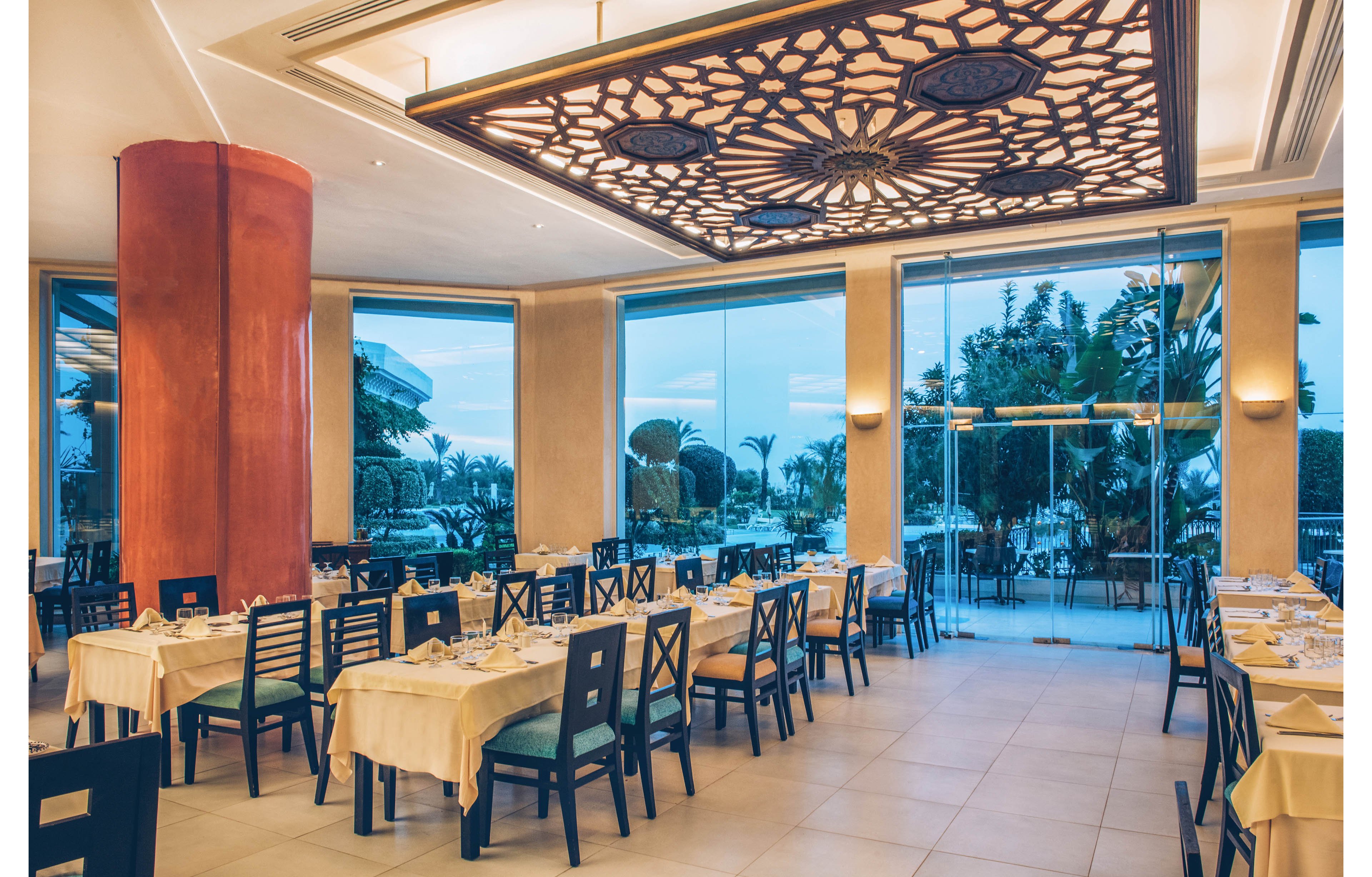 A spacious restaurant dining room is set with neatly arranged tables and dark chairs. Large floor-to-ceiling windows reveal palm trees and a coastal view outside at dusk. A decorative ceiling panel with intricate patterns and warm lighting creates an upscale ambiance. The overall scene feels calm, inviting, and ready for guests.