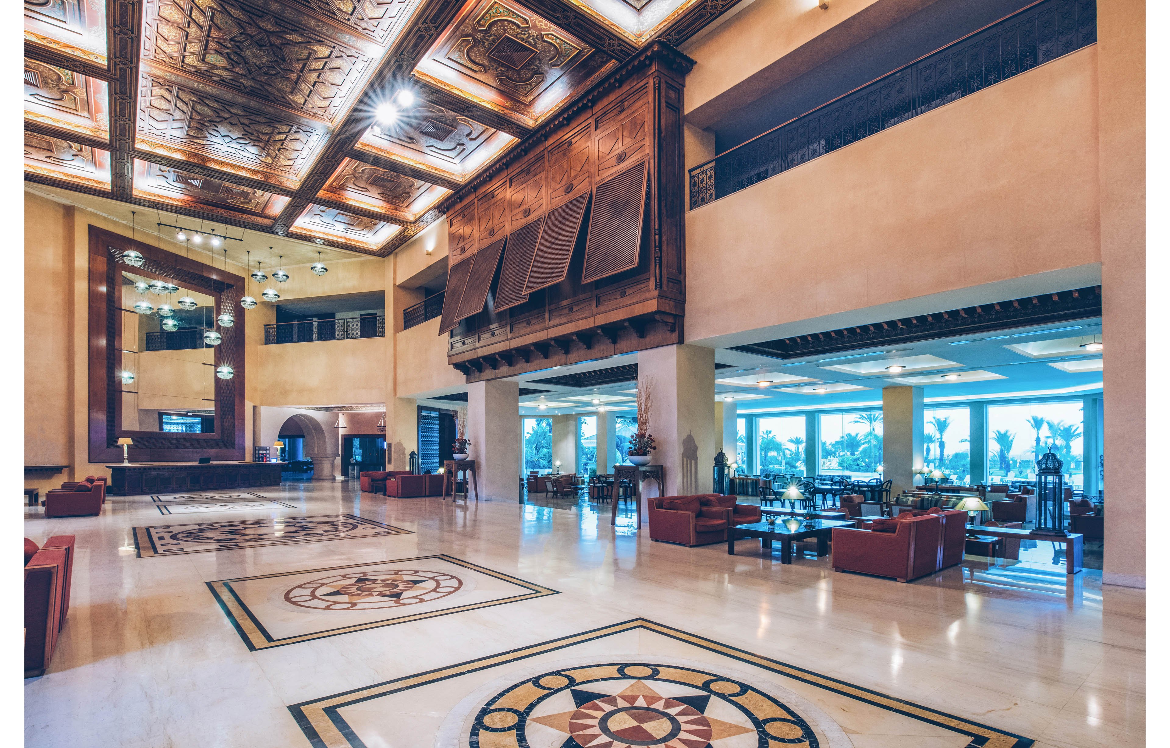 A large, elegant hotel lobby features polished marble floors with decorative geometric inlays. High ceilings with ornate wooden panels and warm lighting create a luxurious atmosphere. Comfortable seating areas with armchairs and sofas are arranged throughout the space. Floor-to-ceiling windows on one side reveal palm trees and an outdoor view, enhancing the open, airy feel.