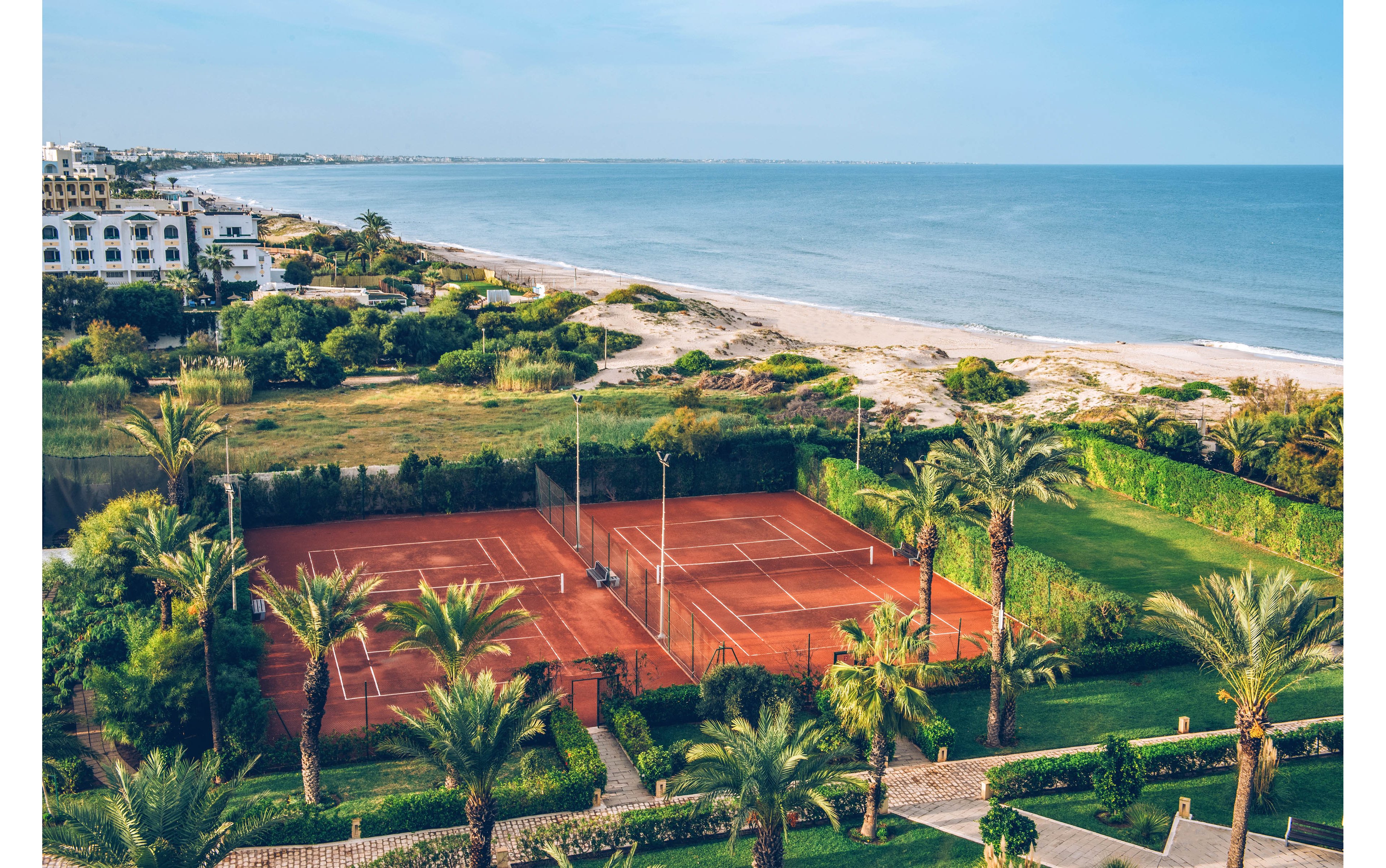 An elevated view shows two red clay tennis courts nestled among lush palm trees near a sandy coastline. The courts are enclosed by green hedges and equipped with tall light poles. Beyond the sports area, grassy fields lead to a wide beach and calm sea. White resort-style buildings line the distant shoreline under a clear blue sky, creating a relaxed vacation atmosphere.