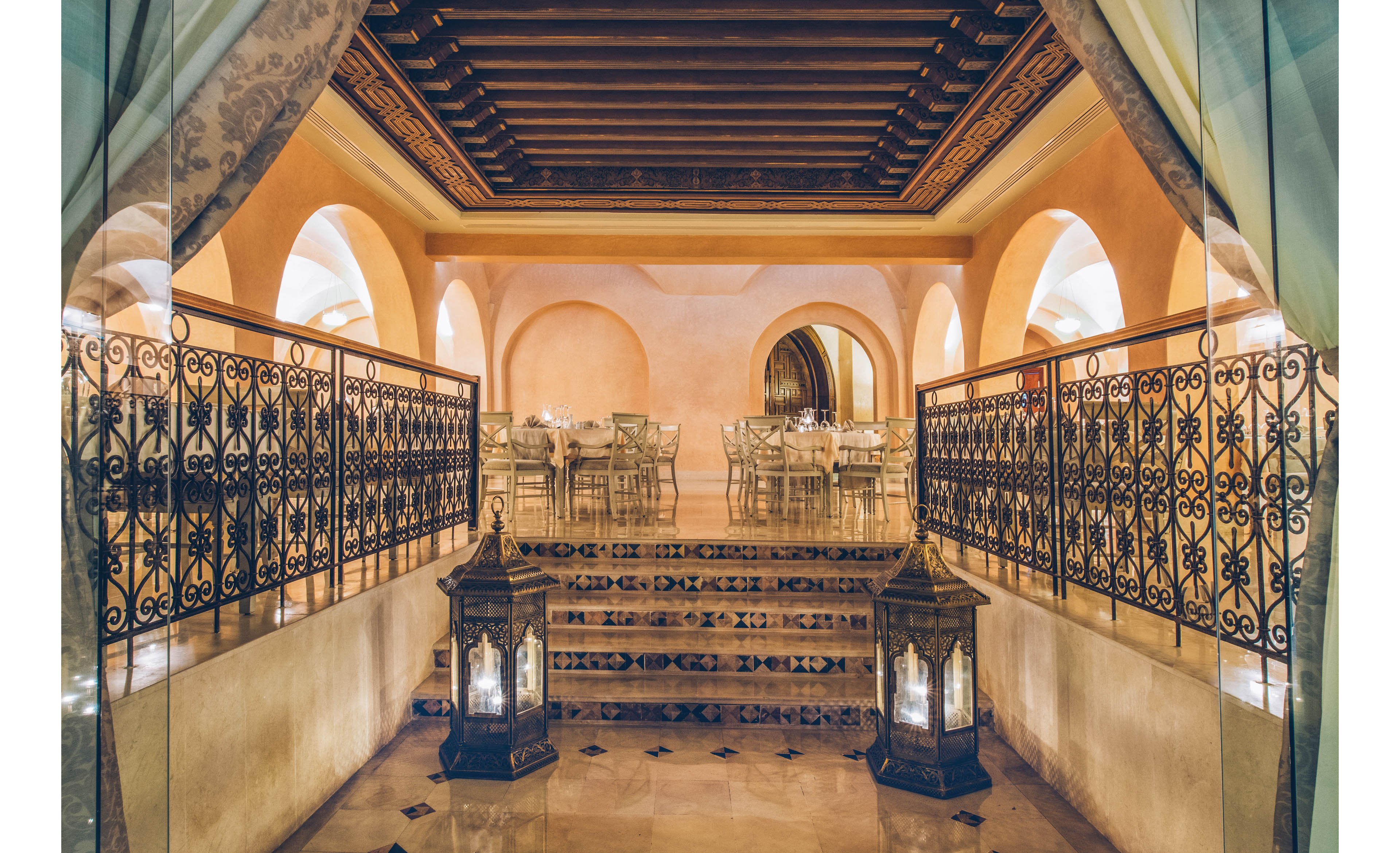 An elegant restaurant interior features warm Mediterranean-style architecture with arched doorways and soft peach-colored walls. Ornate wrought-iron railings frame a short staircase leading up to a dining area set with round tables and chairs. Large decorative lanterns with glowing candles sit at the base of the stairs, adding a romantic ambiance. The polished floor and wooden ceiling beams enhance the luxurious, intimate atmosphere.