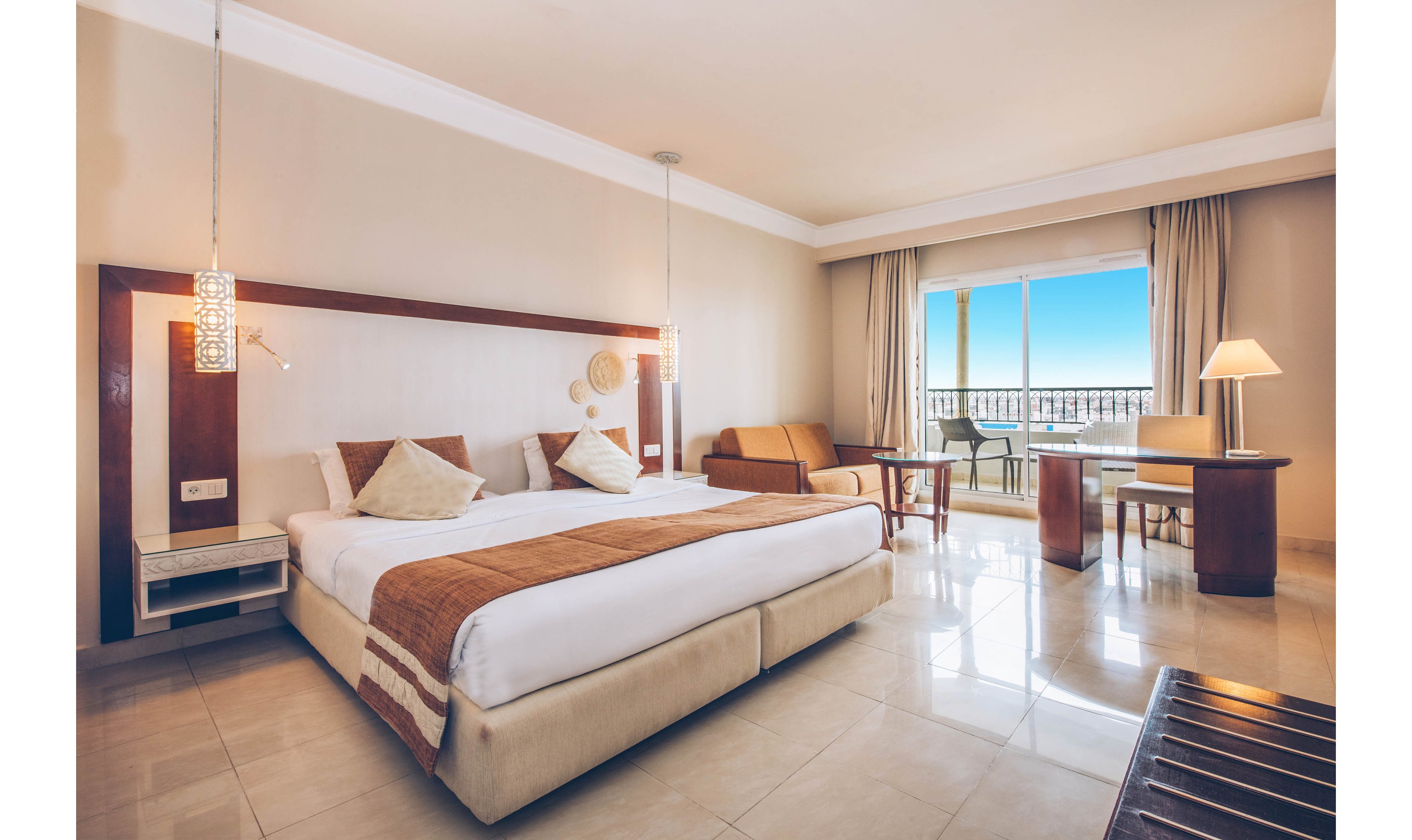 Spacious hotel room featuring a large double bed with neutral bedding and accent pillows. The setting includes a tiled floor, a small sofa, and a work desk with a lamp near the window. Large glass doors open to a balcony with a clear blue sky and distant landscape. Warm lighting and beige tones create a calm, inviting atmosphere suitable for vacation or business travel.