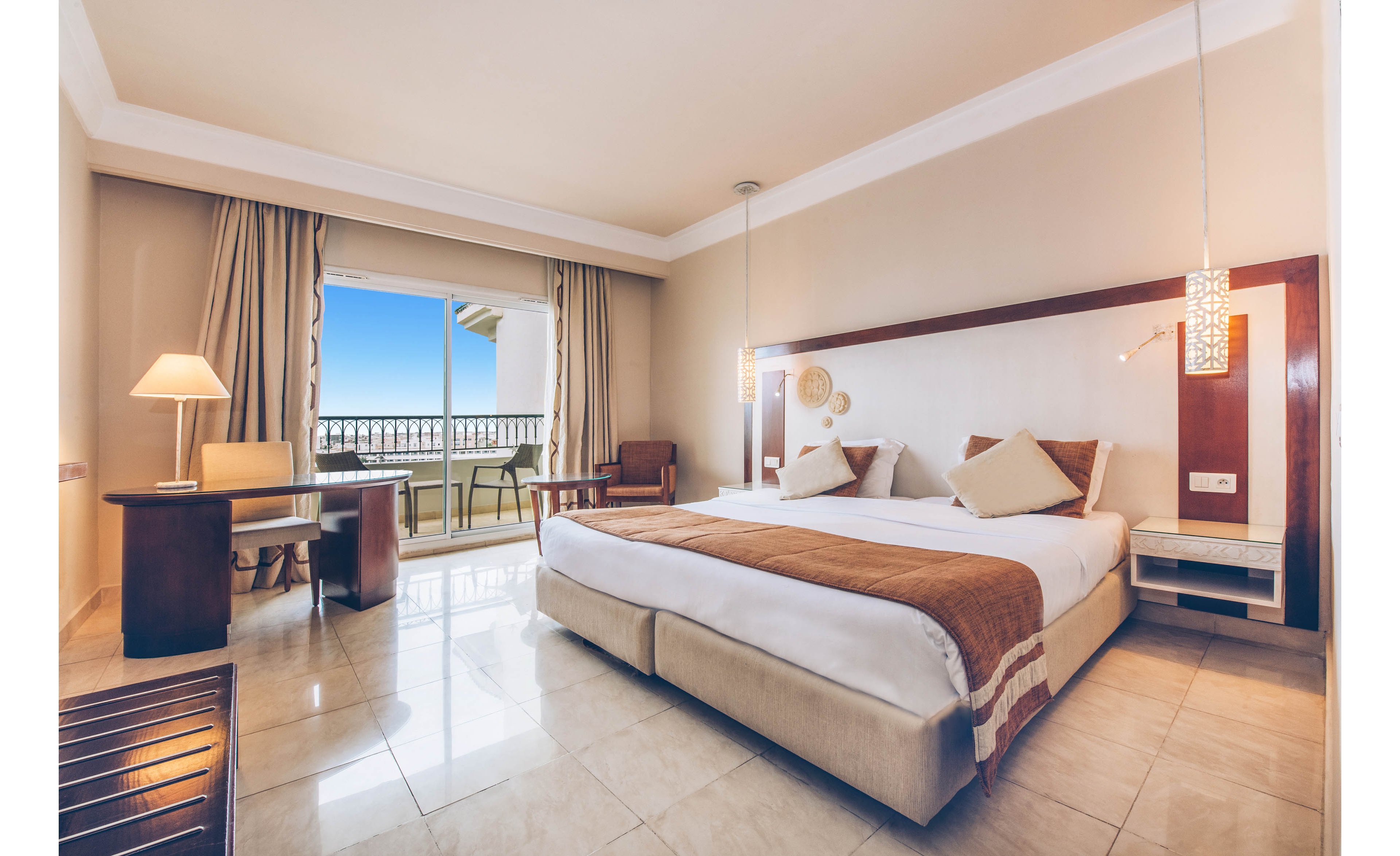 Spacious hotel room featuring a large double bed with white linens and brown accents. The room includes a desk with a lamp, a small seating area, and modern bedside lighting. Large sliding glass doors open to a balcony with chairs and a clear view of the blue sky and sea. Neutral tones and clean lines create a calm, contemporary atmosphere.