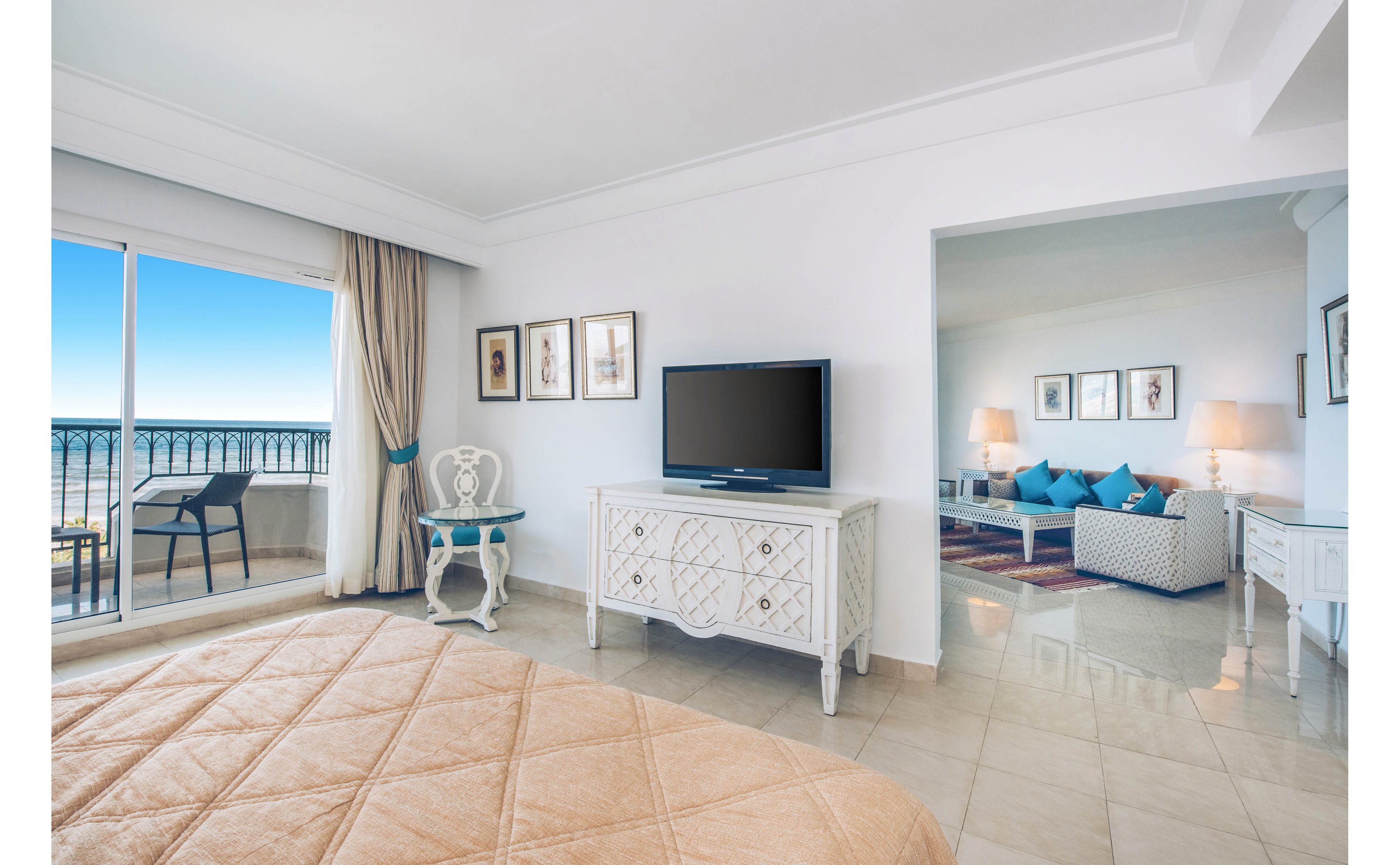 Spacious hotel suite bedroom opens onto a balcony with a clear ocean view. A large bed faces a flat-screen television on a white dresser, with framed artwork on the wall. The adjoining living area features sofas with blue cushions, side tables, and warm table lamps. Light tile flooring and white walls create an airy, minimalist coastal atmosphere.