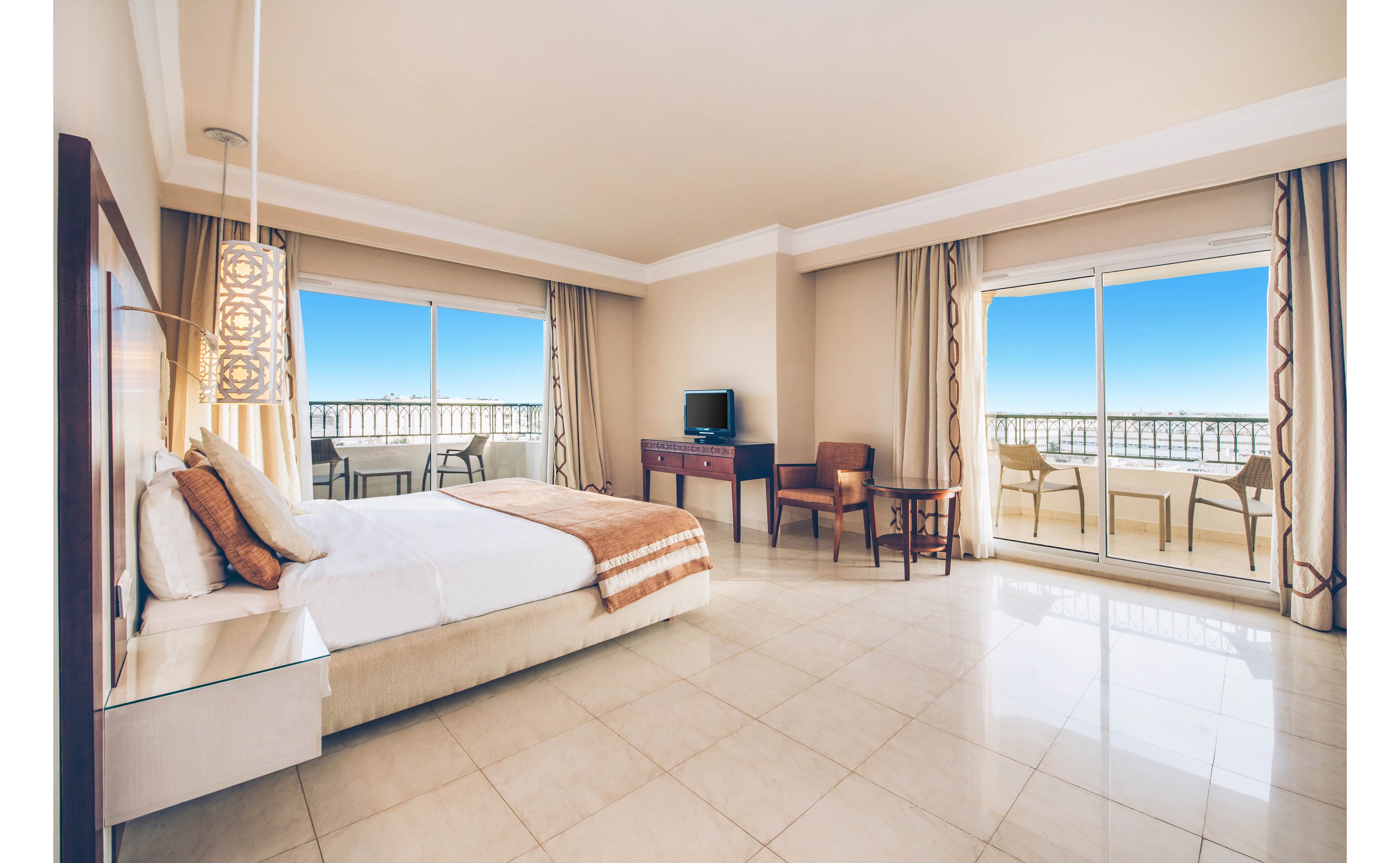 Spacious hotel bedroom with a large bed faces wide glass doors opening to a balcony. The room features tiled floors, neutral beige walls, and contemporary furniture including chairs, a small table, and a dresser with a television. Floor-to-ceiling curtains frame the windows, revealing a clear blue sky and distant water view. Soft lighting and minimal decor create a clean, relaxing atmosphere ideal for vacation or business travel.