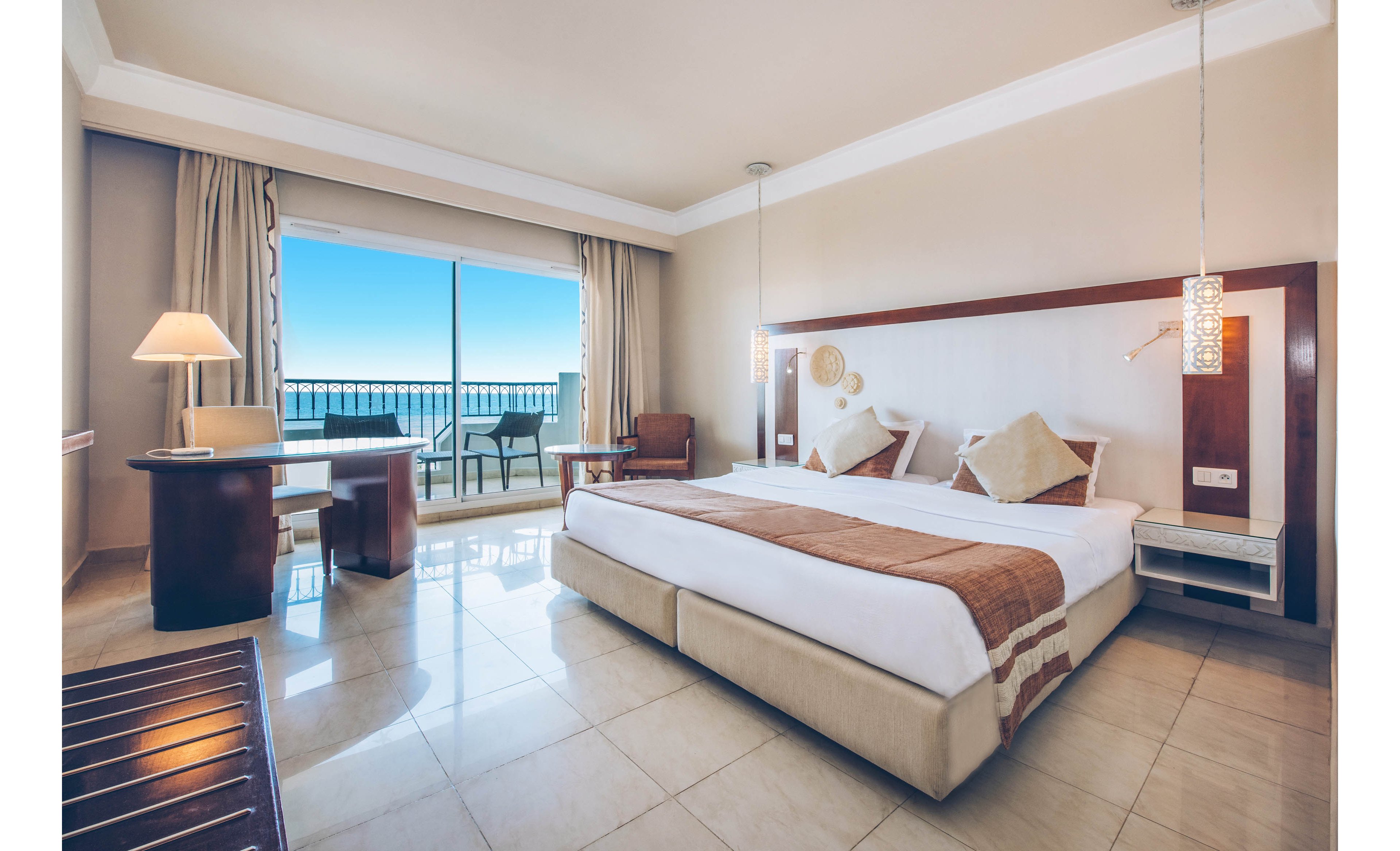 A spacious modern hotel room features a large double bed with neutral bedding and accent pillows. Floor-to-ceiling glass doors open onto a balcony with chairs and a table overlooking a bright blue sea. The room includes a wooden desk with a lamp, an armchair, and contemporary pendant lighting. Light tile flooring and beige walls create an airy, minimalist atmosphere ideal for vacation or business travel.