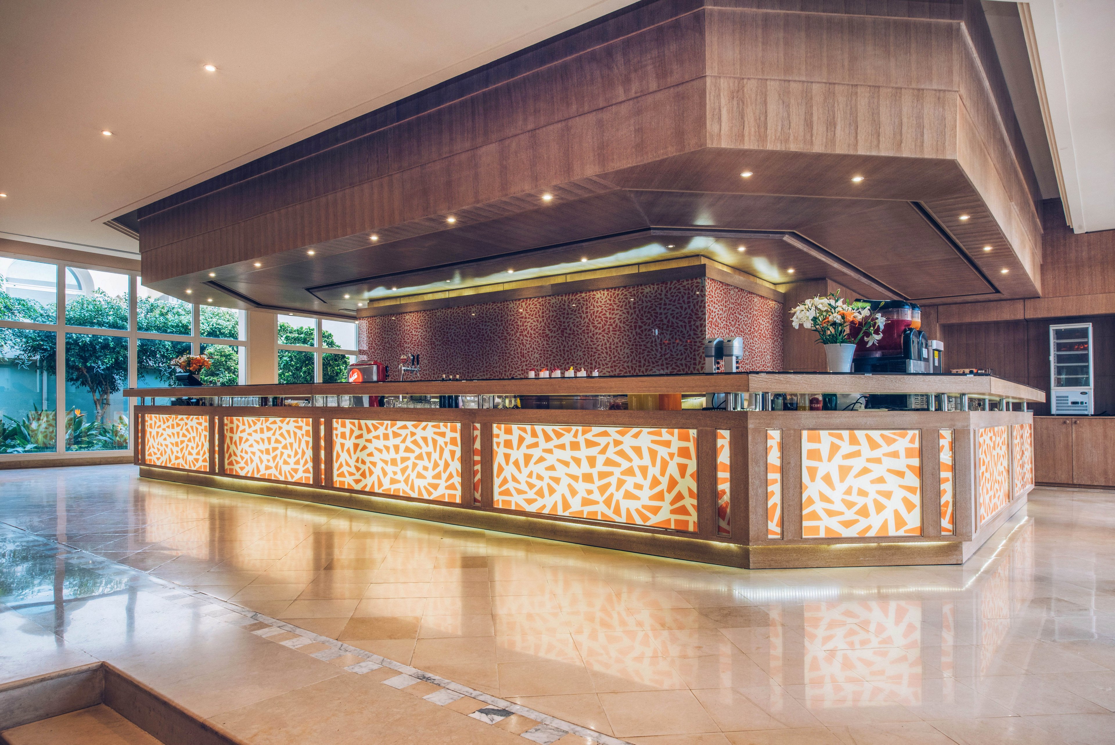 A spacious, contemporary bar counter dominates the interior of a hotel or resort lobby. The bar features illuminated geometric panels in warm orange and white tones, framed by sleek wooden finishes. Recessed ceiling lights and under-counter lighting create a cozy, upscale atmosphere. Large windows in the background reveal greenery outside, adding a touch of nature to the refined indoor setting.