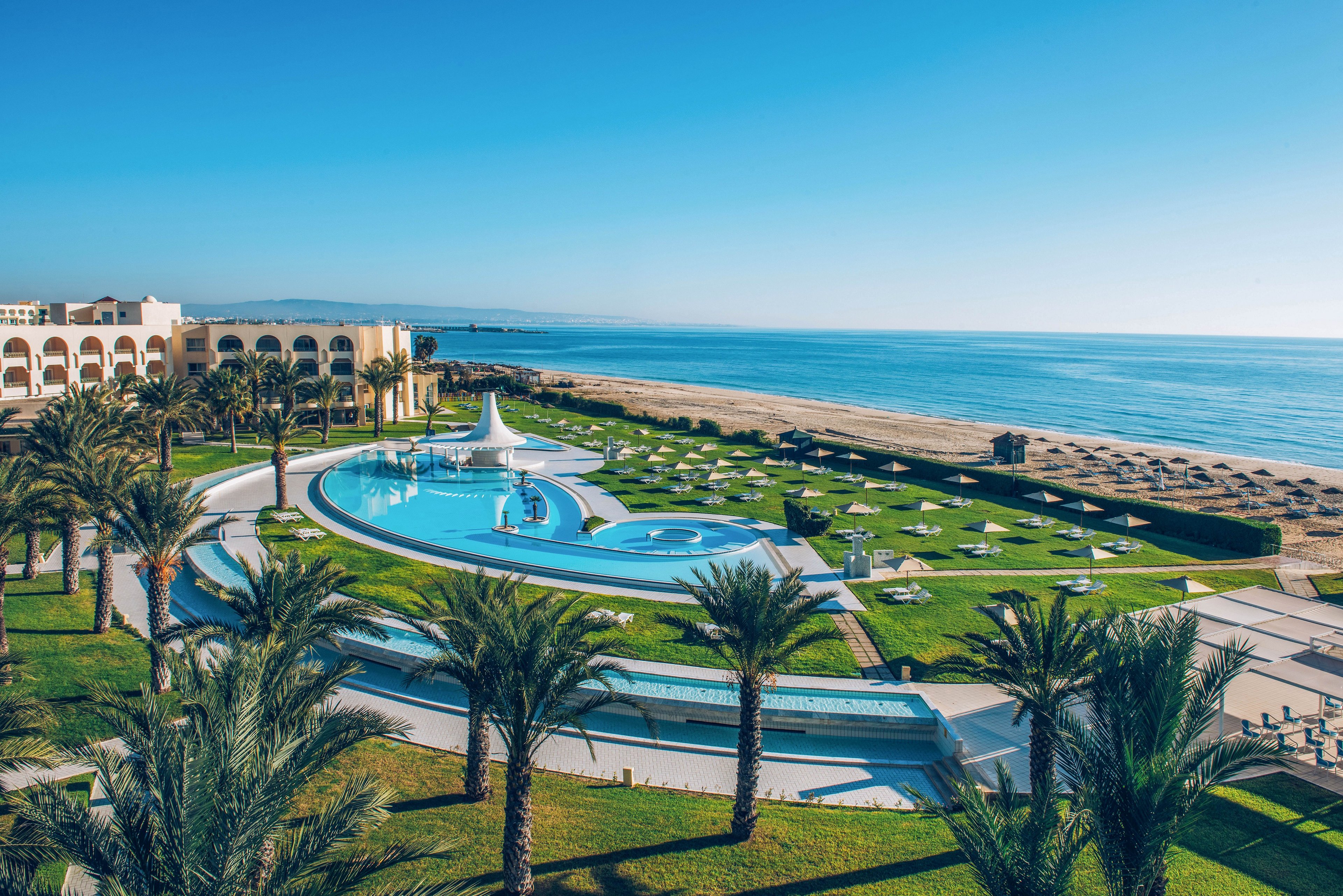A luxurious seaside resort features a large curved swimming pool surrounded by palm trees and manicured green lawns. Sun loungers and umbrellas are neatly arranged across the grass and along the sandy beach. The calm blue sea stretches to the horizon under a clear sky, creating a tranquil vacation atmosphere. Elegant white buildings with arches frame the scene, emphasizing the upscale coastal setting.