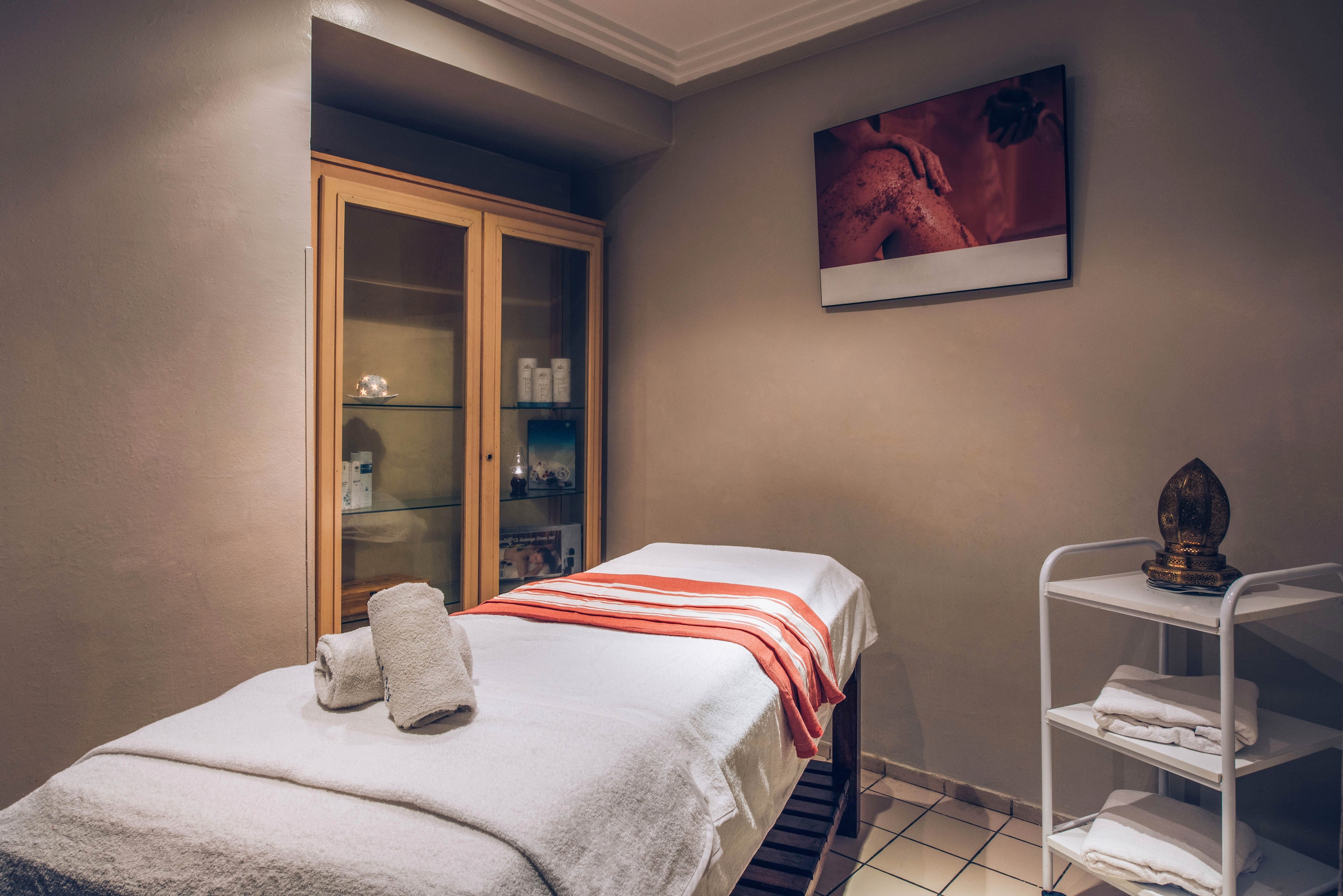 A neatly prepared massage table covered with white linens and a red striped towel stands in a softly lit spa room. Rolled towels are placed at the head of the table, ready for a treatment. A glass-front cabinet in the background holds skincare and wellness products. A small trolley with folded towels and a decorative object adds to the serene, professional atmosphere.