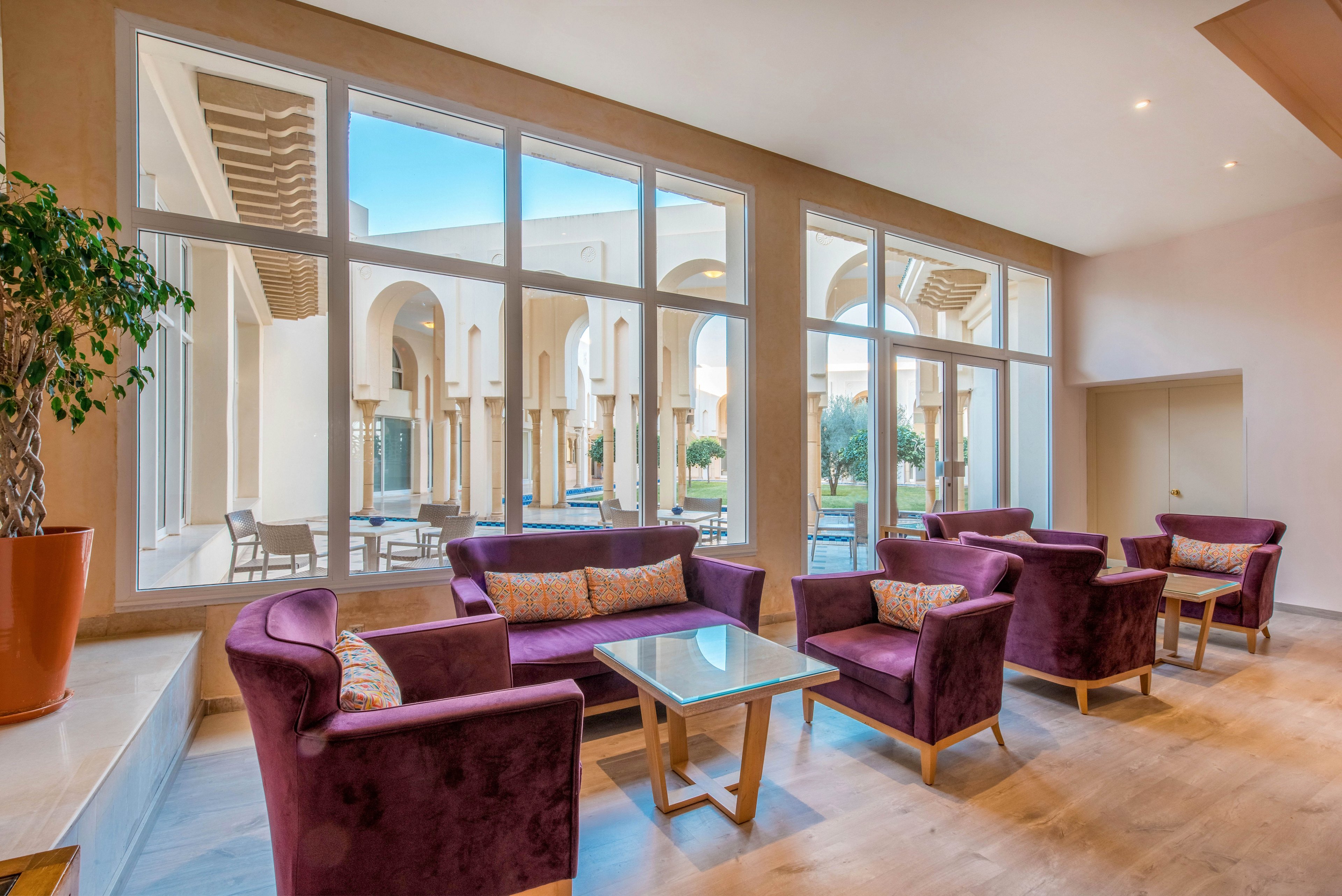 A contemporary lounge area features plush purple armchairs and sofas arranged around glass-topped coffee tables. Large floor-to-ceiling windows reveal an outdoor courtyard with arches and columns in a warm, Mediterranean-inspired style. Natural light floods the interior, highlighting the light wood flooring and neutral walls. The space feels airy and inviting, suitable for relaxing, socializing, or informal meetings.