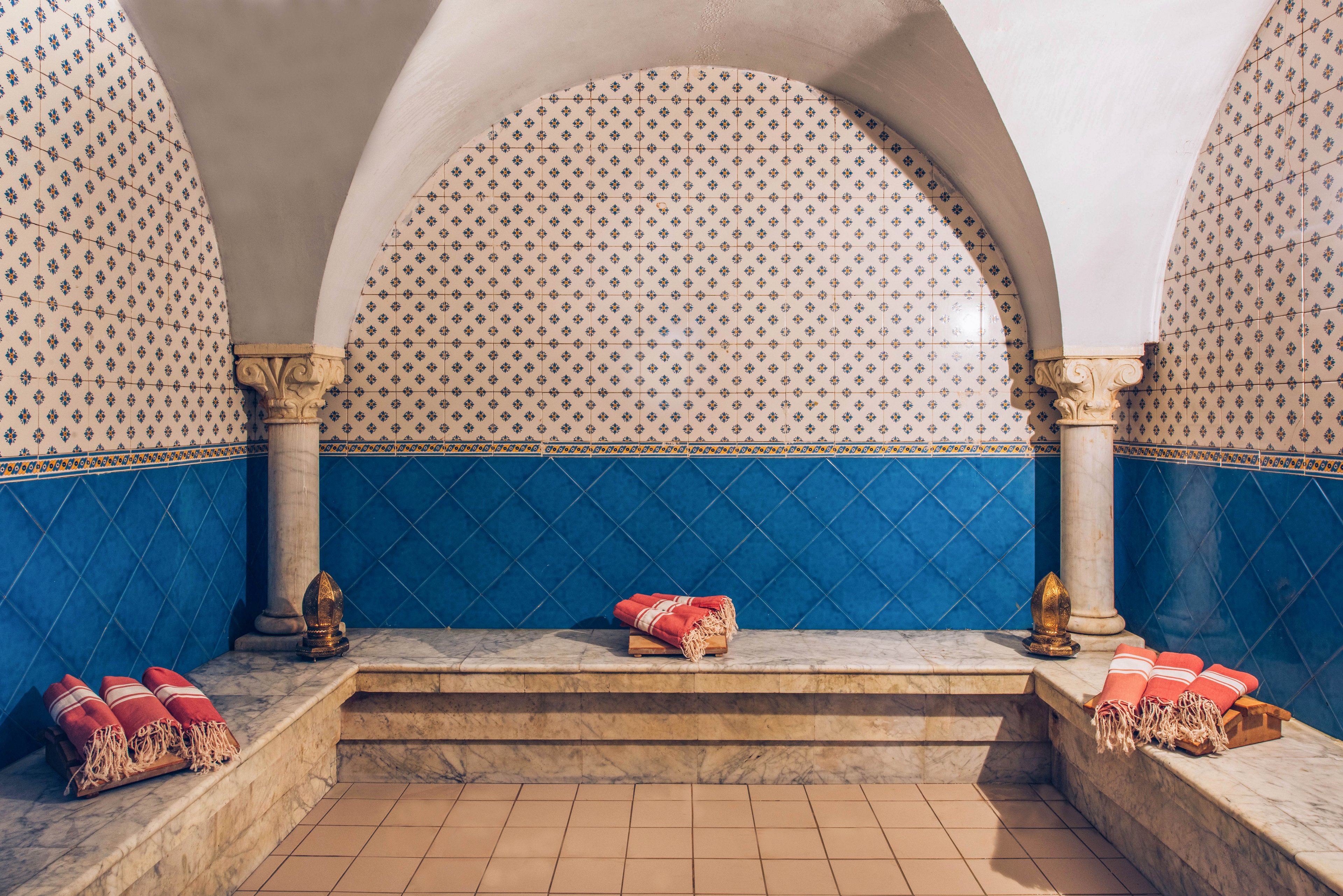 A traditional hammam interior features arched ceilings and patterned wall tiles. Blue diamond tiles line the lower walls, while smaller geometric tiles cover the upper section and arch. Stone benches run along the sides and back, topped with neatly folded striped towels. Warm lighting and decorative metal lanterns enhance the serene, spa-like atmosphere.