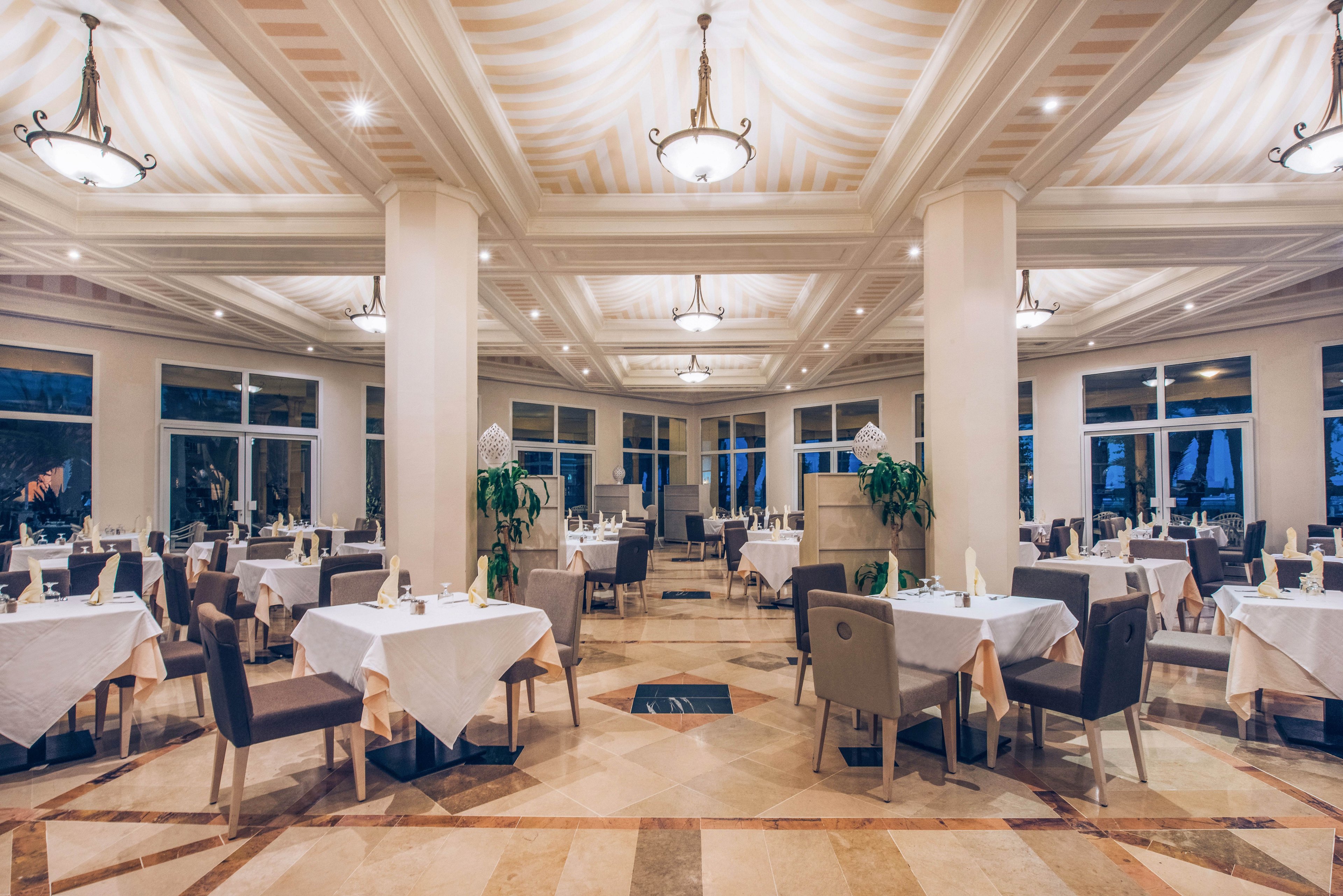 Spacious restaurant dining room with neatly arranged tables and chairs is shown empty and ready for guests. White tablecloths, folded napkins, and simple place settings create a refined atmosphere. Large windows line the walls, revealing a dark evening sky outside. Warm ceiling lights and decorative columns add to the sophisticated, tranquil ambiance.