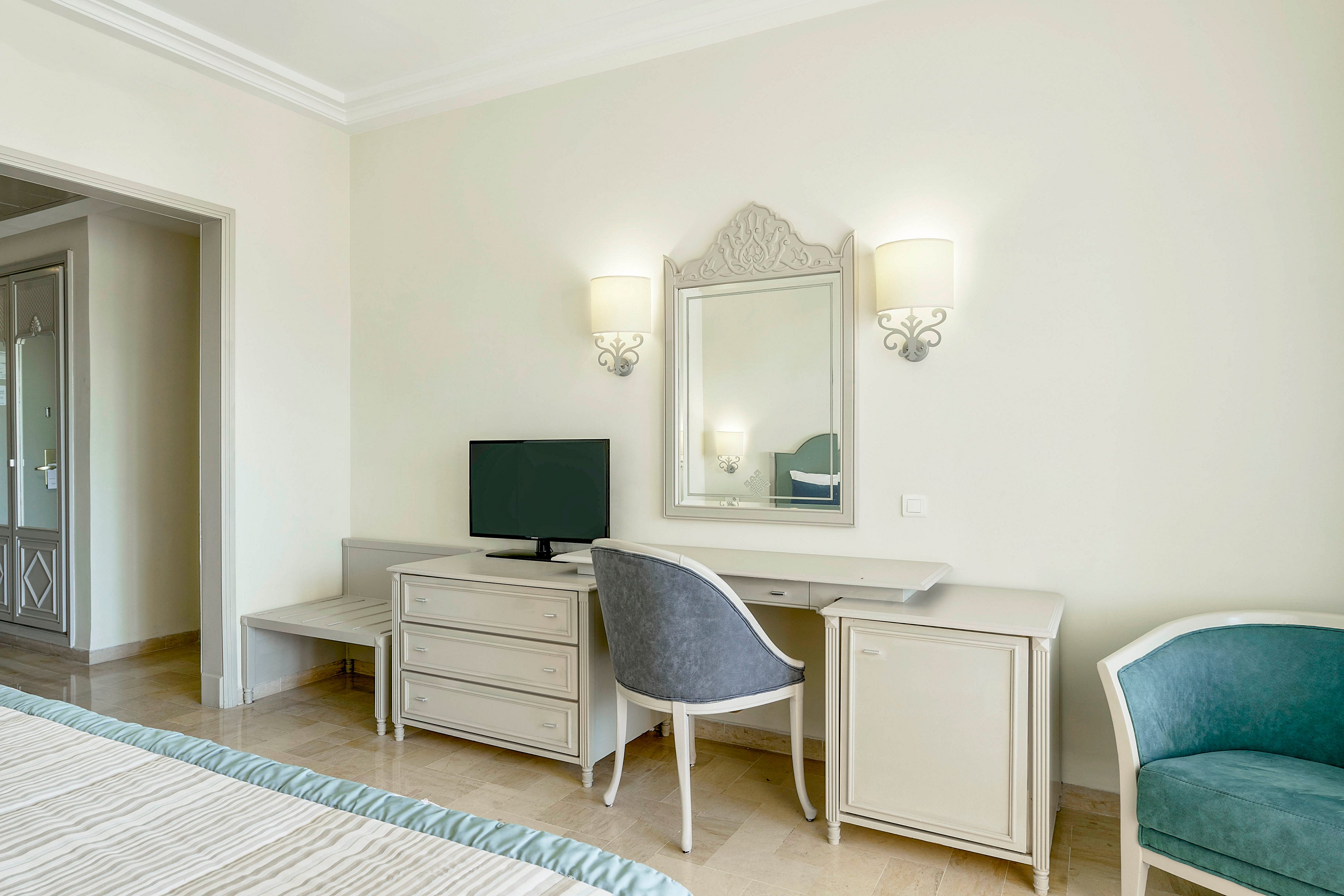 A bright hotel room features a classic desk area with a framed mirror and two wall sconces. A small flat-screen television sits on the desk beside a cushioned chair. Neutral cream walls and light wood furniture create an airy, minimalist atmosphere. A teal upholstered armchair and bed edge add subtle color accents to the space.