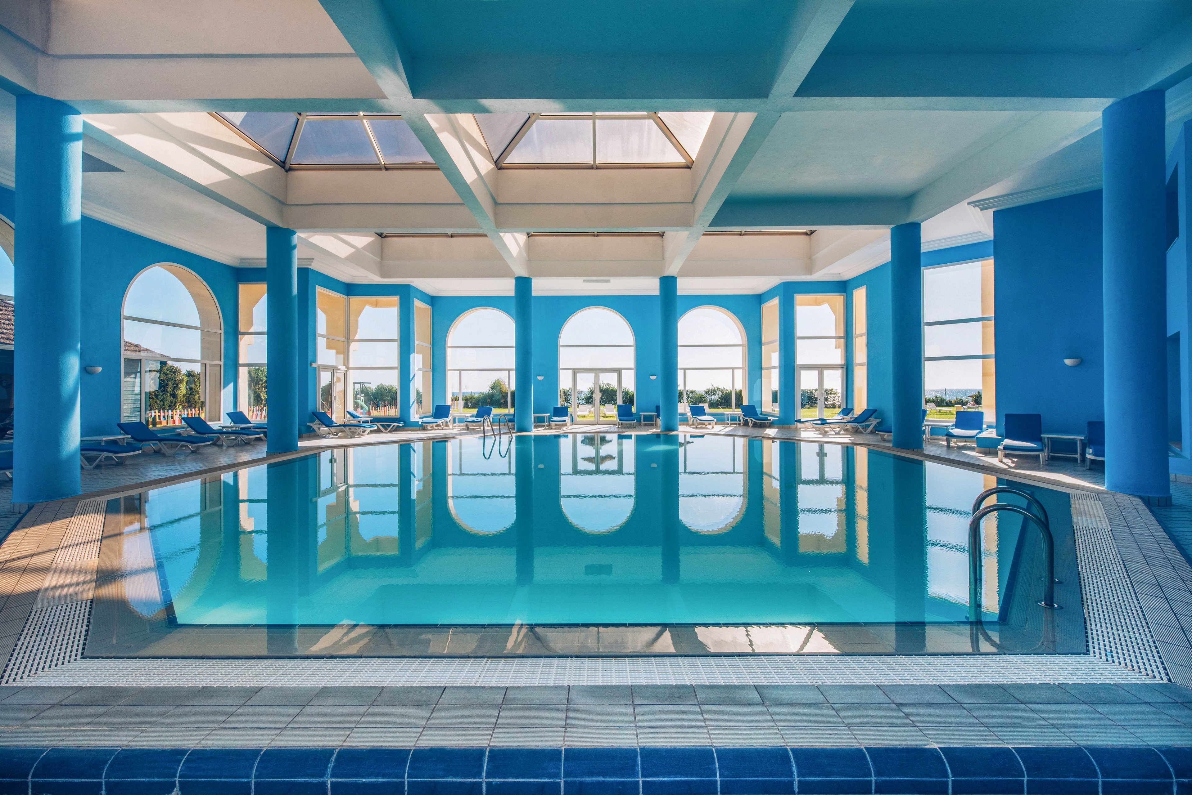 Spacious indoor swimming pool in a resort-style building with bright blue walls and columns. Large arched windows and skylights let in abundant natural light and reveal an outdoor landscape. Sun loungers line the sides of the pool, suggesting a relaxing vacation or spa environment. The calm water reflects the architecture, creating a symmetrical and tranquil scene.