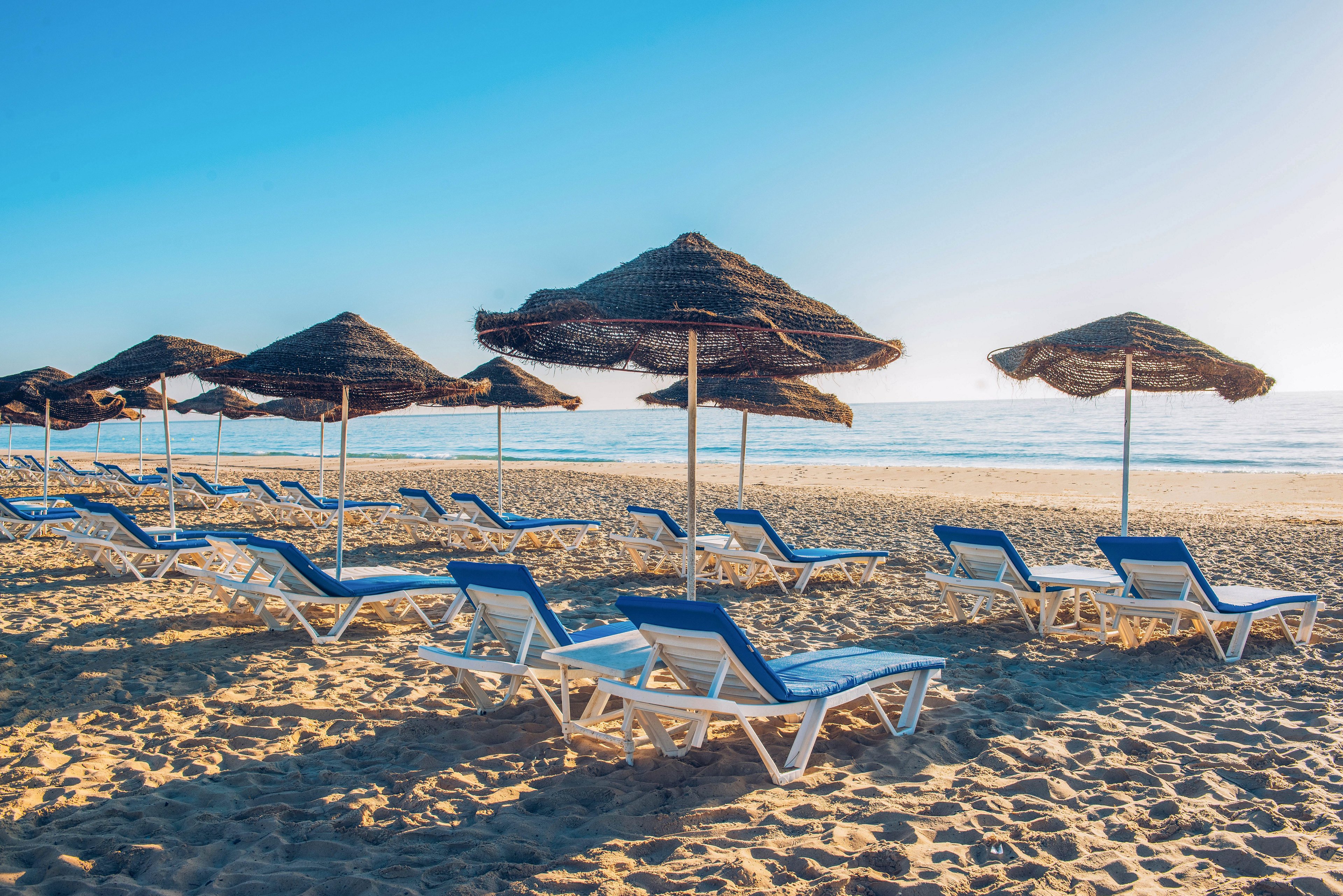 A row of white sun loungers with blue cushions sits on a sandy beach under straw umbrellas. The scene overlooks a calm sea with gentle waves meeting the shoreline. The sky is clear and bright blue, suggesting a sunny, warm day. The absence of people creates a peaceful, relaxing vacation atmosphere.
