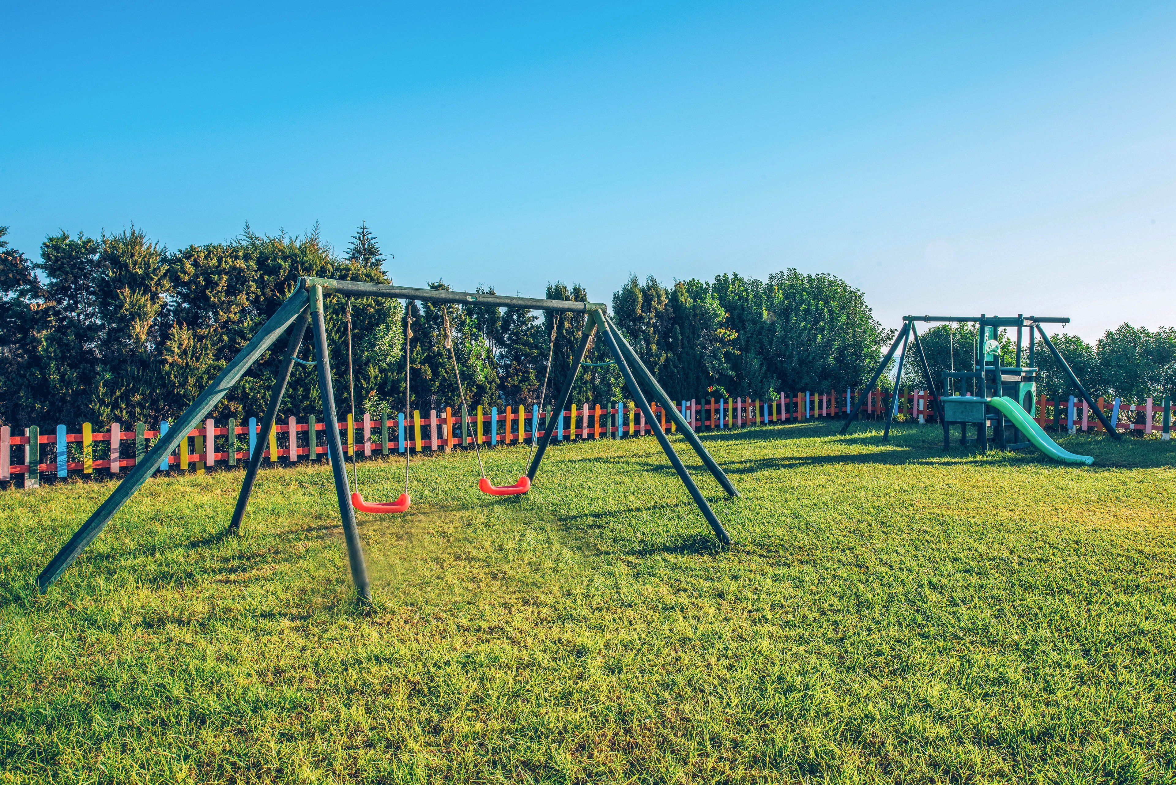 An outdoor playground stands empty under a clear blue sky. Two red swings hang from a metal frame on a wide grassy field. In the background, a play structure with a slide is visible near a colorful fence and trees. The bright daylight and vivid colors create a cheerful, inviting atmosphere.