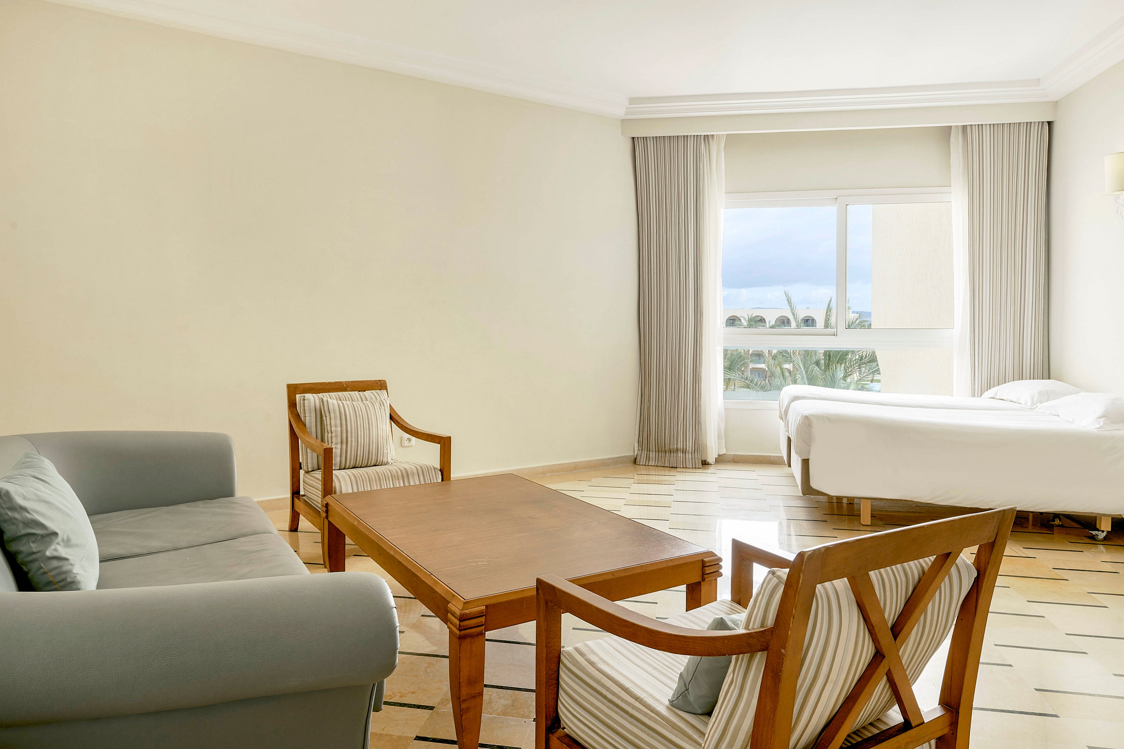 A spacious hotel room features a simple seating area with a sofa, two armchairs, and a wooden coffee table. In the background, a single bed is positioned near a large window with light curtains. Natural daylight fills the room, highlighting the neutral color palette of beige walls and tiled floor. The overall atmosphere is calm, clean, and minimal, suitable for relaxation or quiet work.