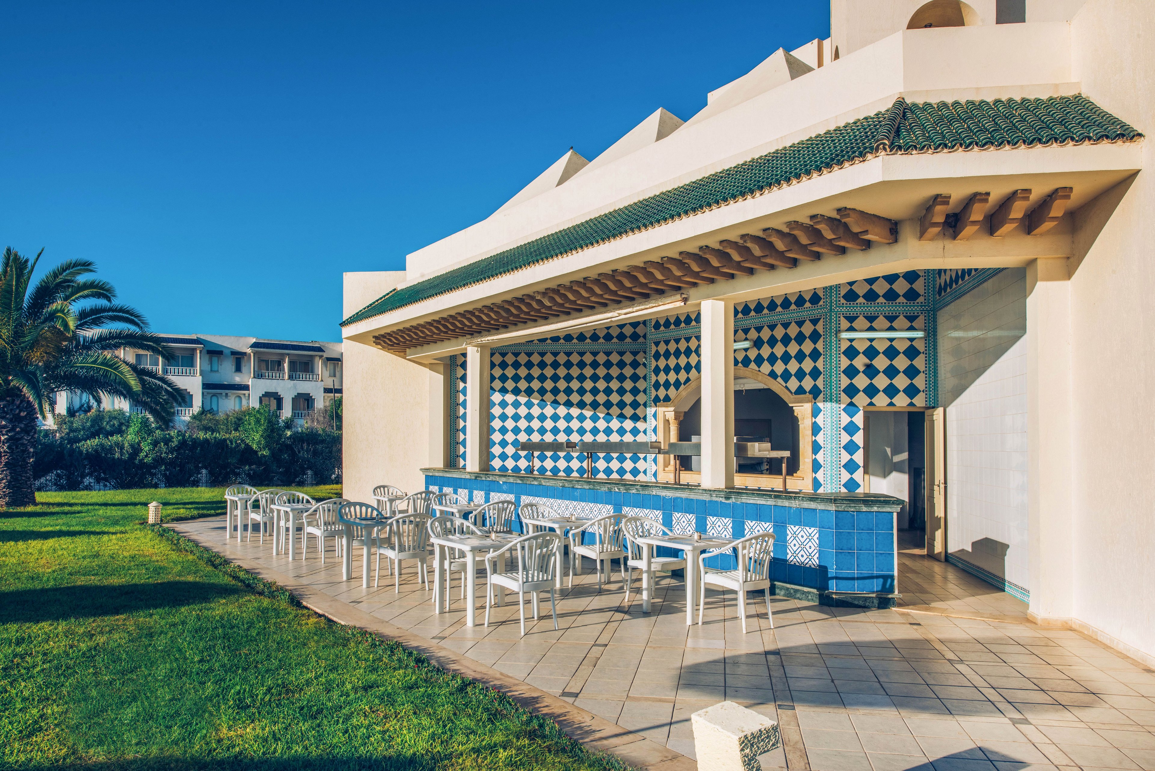 An open-air bar with blue and white geometric tiles stands beside a sunlit terrace. White plastic tables and chairs are neatly arranged on the paved patio, ready for guests. A palm tree and manicured green lawn frame the scene, with additional resort buildings visible in the background. Clear blue sky and strong sunlight create a bright, inviting holiday atmosphere.