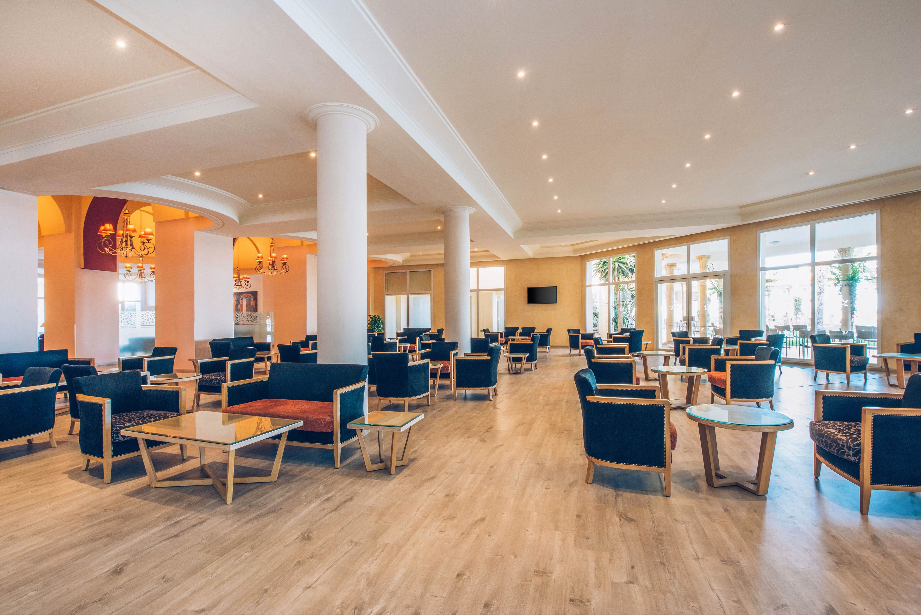 A large, modern hotel lobby lounge features numerous armchairs and small glass-topped tables arranged in neat rows. Light wood flooring and white columns create an airy, open atmosphere. Recessed ceiling lights and large windows provide bright, even illumination throughout the space. The lounge appears quiet and empty, emphasizing its clean, contemporary design and comfortable seating areas.