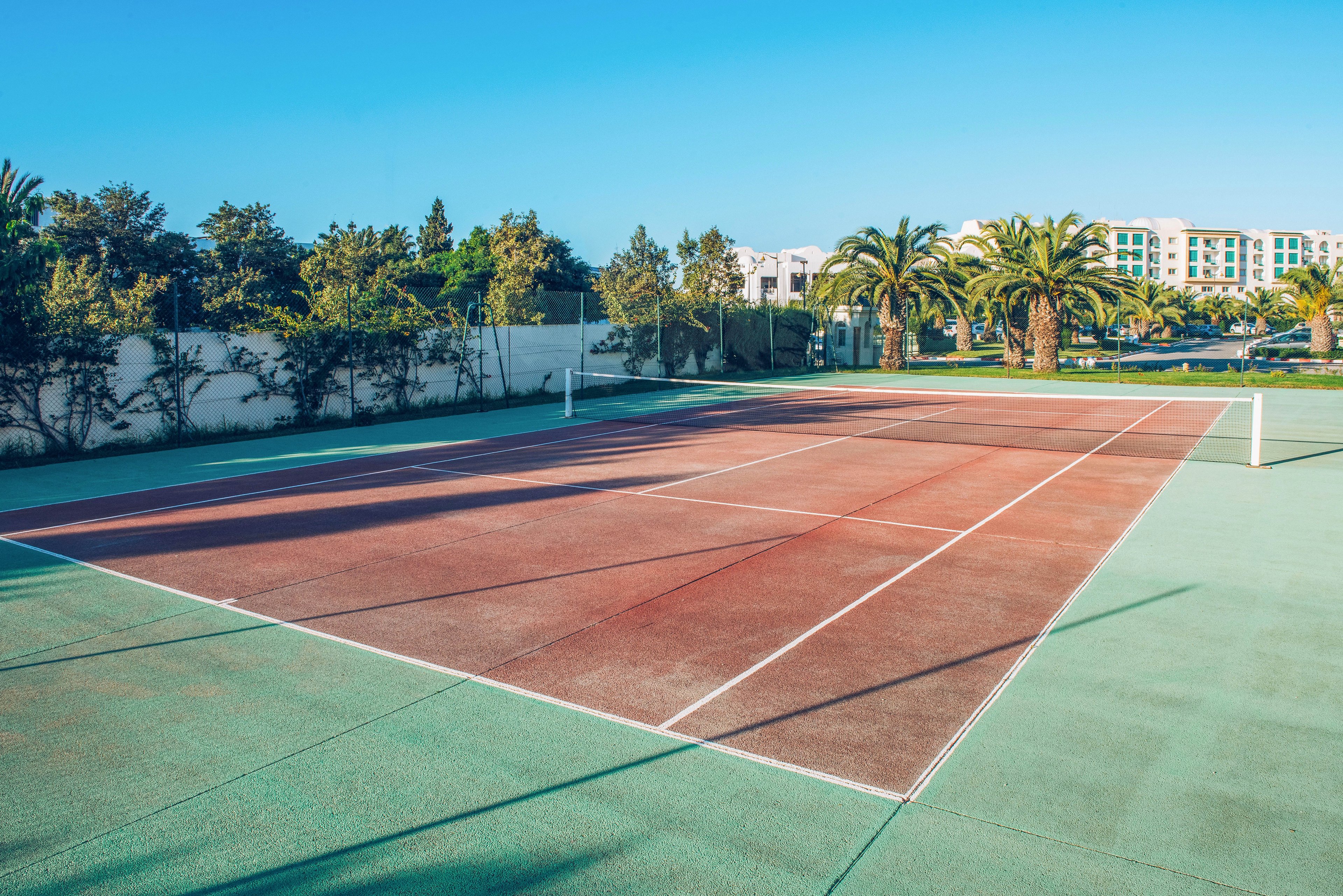 An empty outdoor tennis court is shown under bright daylight, with clear white lines on a red and green surface. Palm trees and lush greenery border the court, creating a relaxed resort atmosphere. In the background, low-rise modern buildings are visible against a clear blue sky. The scene feels calm and inviting, suggesting leisure and vacation sports activities.