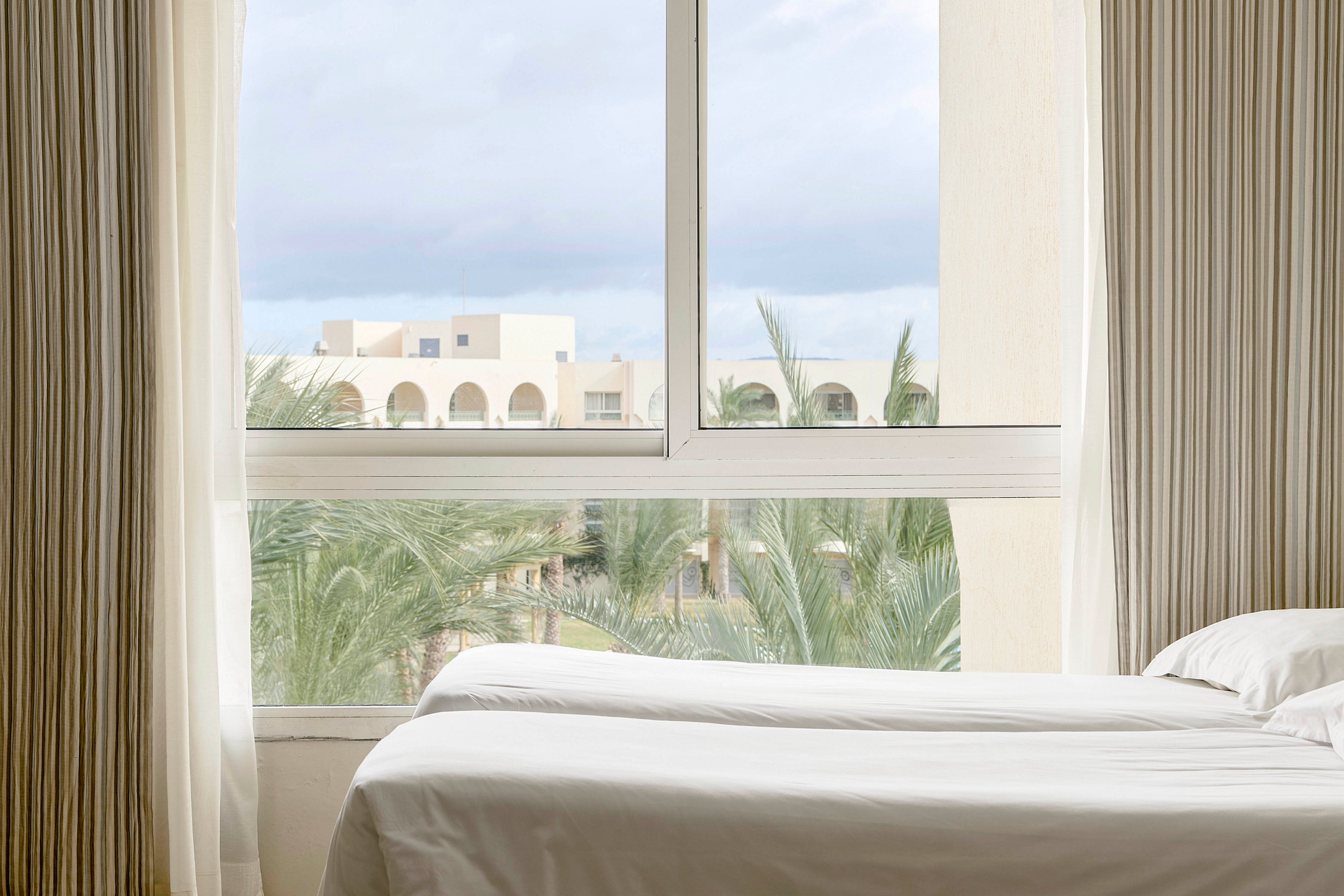 A neatly made bed sits beside a large window in a bright hotel room. Through the window, a resort complex with arched balconies and light-colored buildings is visible. Palm trees fill the foreground, adding a tropical feel to the scene. Soft curtains frame the window, creating a calm and relaxing atmosphere.