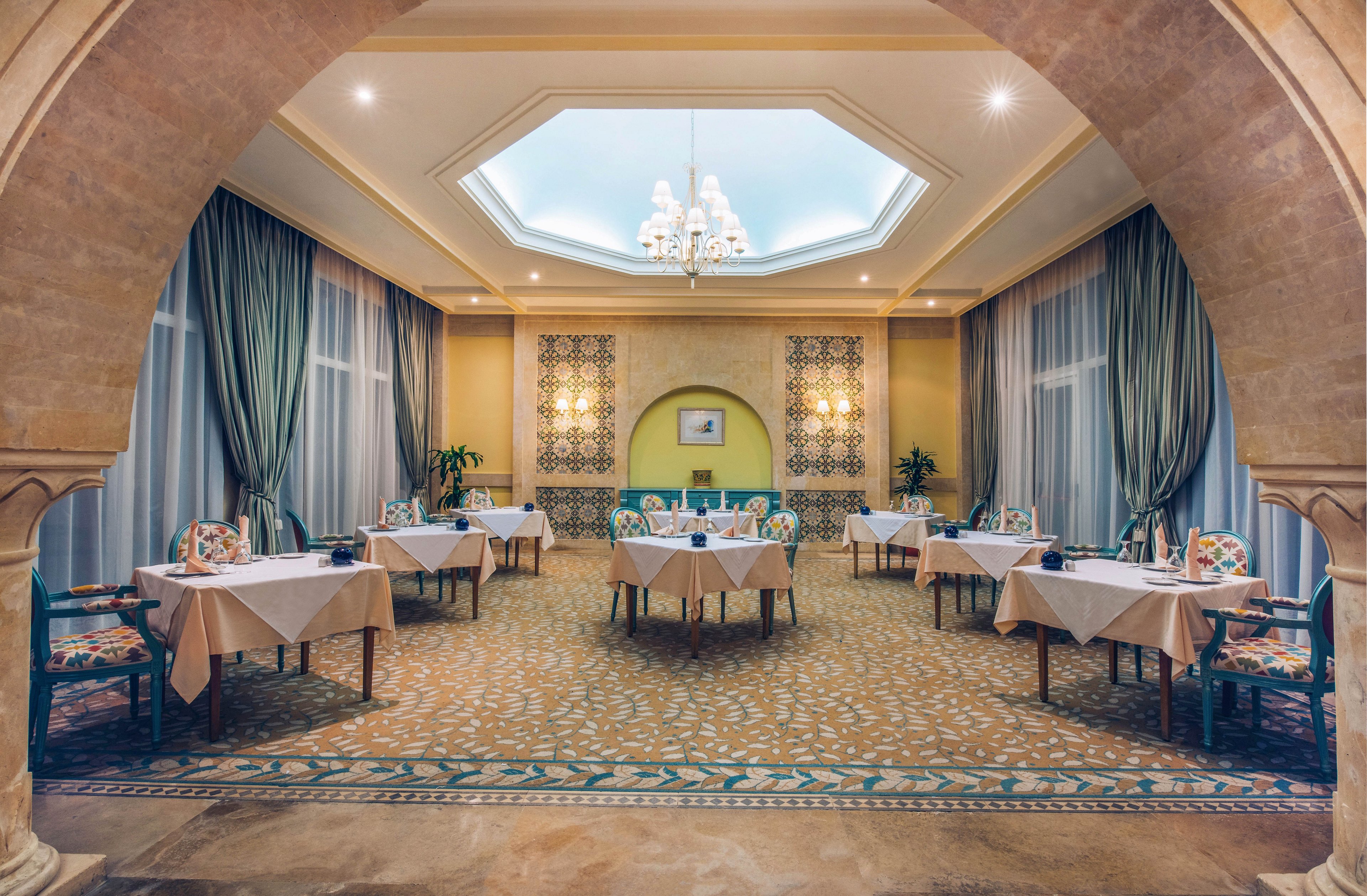 An upscale restaurant dining room is arranged with neatly set tables and upholstered chairs. The space features an arched entrance framing the view toward a central alcove with decorative wall patterns and artwork. Tall windows with flowing curtains line the walls, and a chandelier hangs from a recessed ceiling. Warm lighting and soft colors create a calm, refined atmosphere in the empty room.