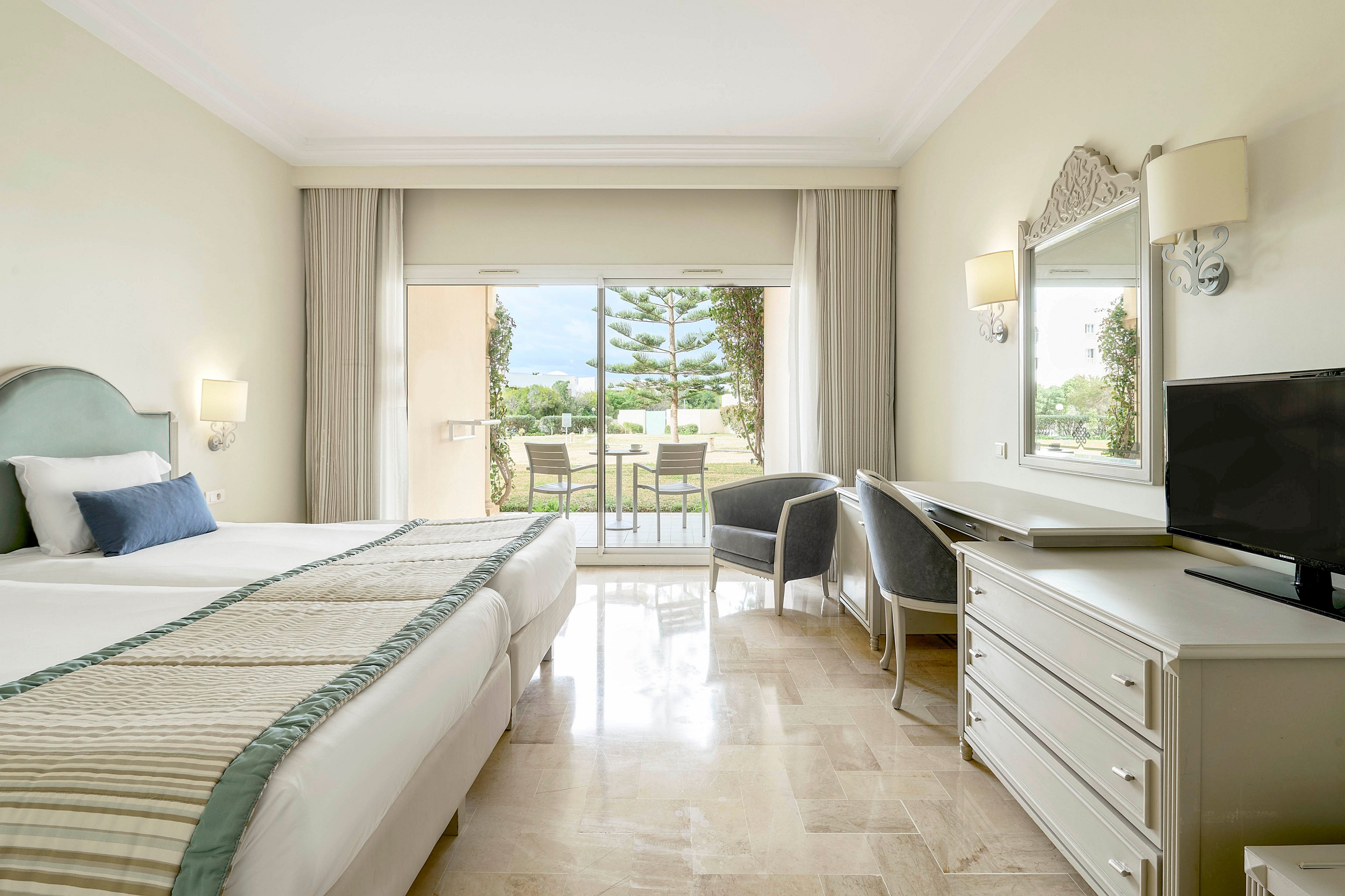 A spacious hotel bedroom features a large neatly made bed with striped bedding and accent pillows. The room includes a desk with a flat-screen television, a mirror, and a comfortable chair. Neutral walls and light marble flooring create an airy, minimalist atmosphere. Large glass doors open to a balcony with outdoor seating and a view of greenery and trees beyond.