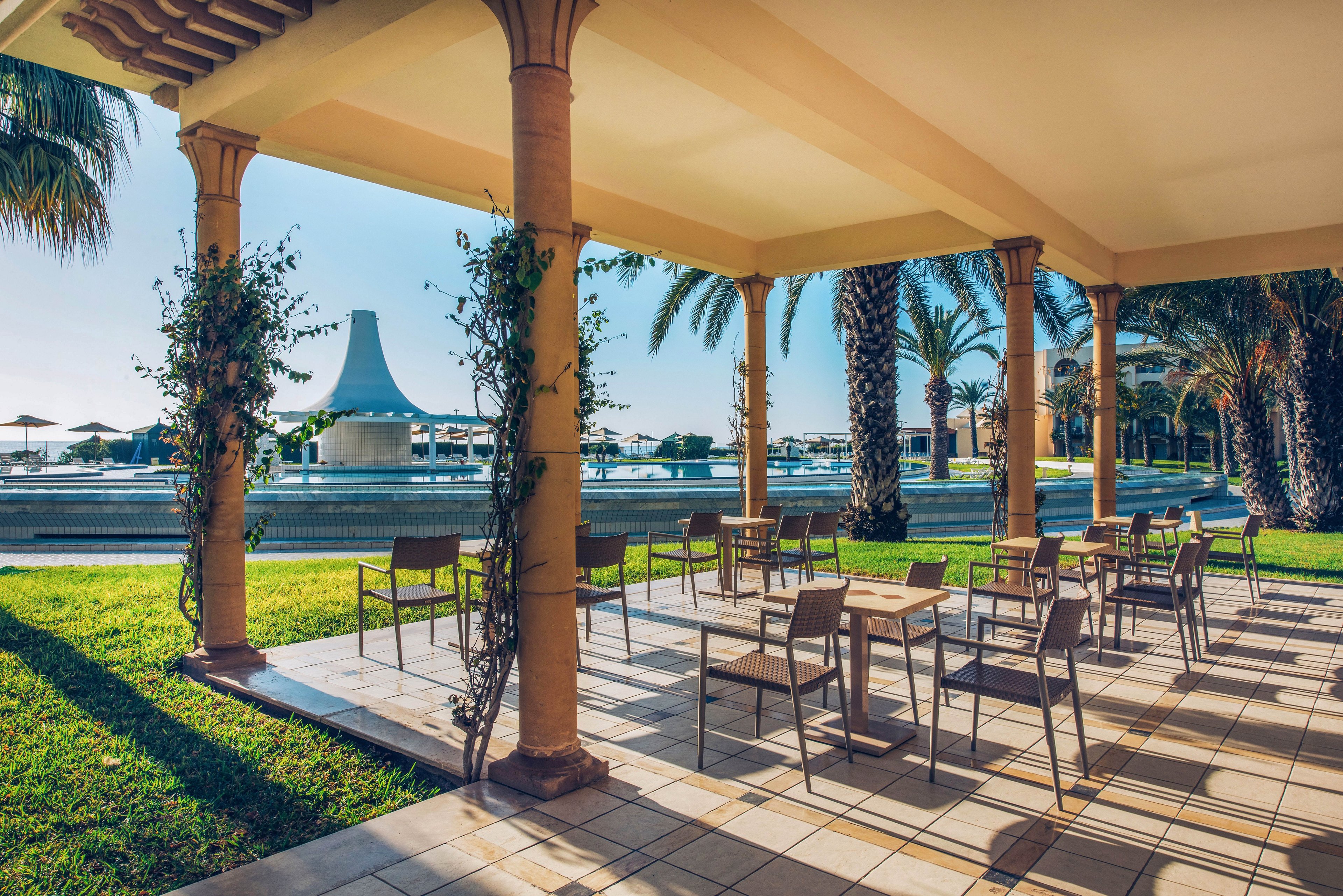 An open-air terrace with several empty tables and chairs overlooks a large resort swimming pool. Tall columns wrapped with climbing plants frame the view toward palm trees and sun loungers in the distance. Bright morning or late afternoon sunlight casts long shadows across the tiled floor, creating a warm and inviting atmosphere. The scene suggests a tranquil vacation setting ready for outdoor dining or relaxation.