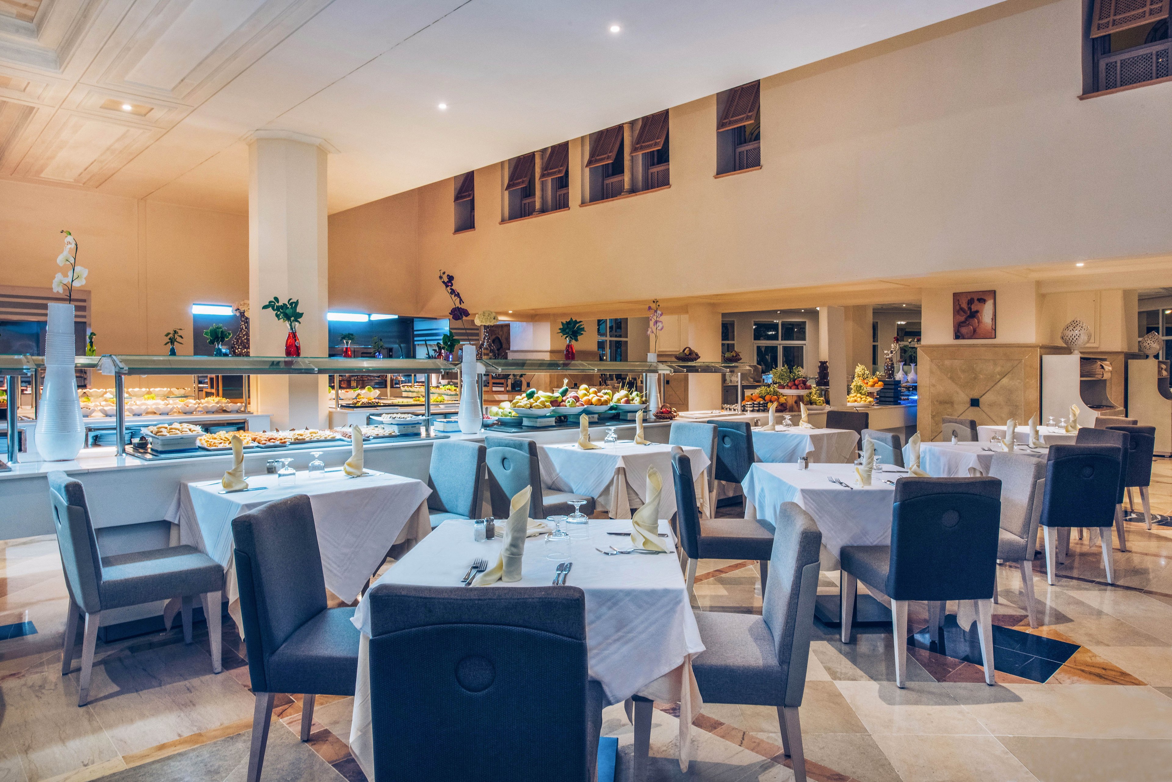 A spacious restaurant dining area features neatly arranged tables covered with white tablecloths and set with folded napkins and glassware. Modern dark chairs surround each table, creating a refined contrast against the light tiled floor. In the background, a brightly lit buffet counter showcases a wide variety of dishes and desserts. Large windows and warm interior lighting contribute to an inviting, upscale atmosphere suitable for evening dining.