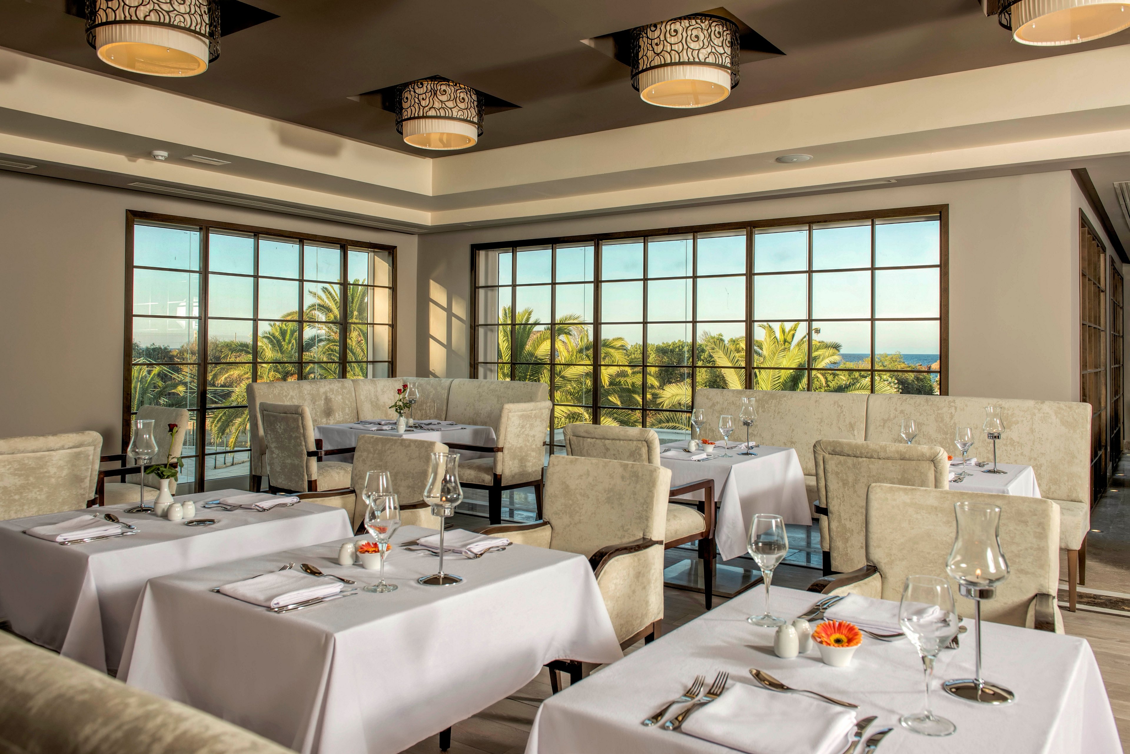 Upscale restaurant dining room with neatly set tables and upholstered chairs overlooks a scenic coastal landscape. Large floor-to-ceiling windows reveal palm trees and a distant ocean horizon under clear skies. White tablecloths, folded napkins, wine glasses, and small floral centerpieces create a refined, formal atmosphere. Warm ceiling lights and neutral decor contribute to a calm, sophisticated ambiance suitable for fine dining.