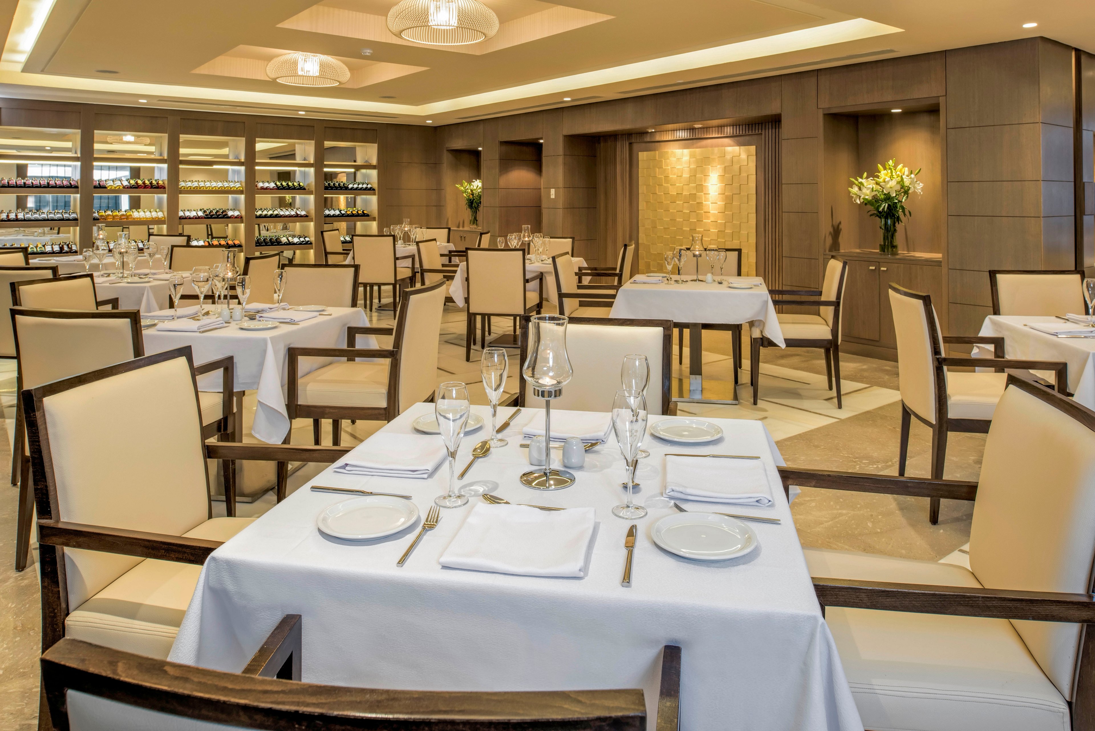A sophisticated restaurant dining room is set with neatly arranged tables covered in white tablecloths. Wooden chairs with light upholstery surround each table, which is prepared with plates, glasses, and folded napkins. Warm recessed lighting and modern ceiling fixtures create a soft, inviting ambiance. Shelves of wine bottles and decorative flower arrangements enhance the upscale, contemporary atmosphere.