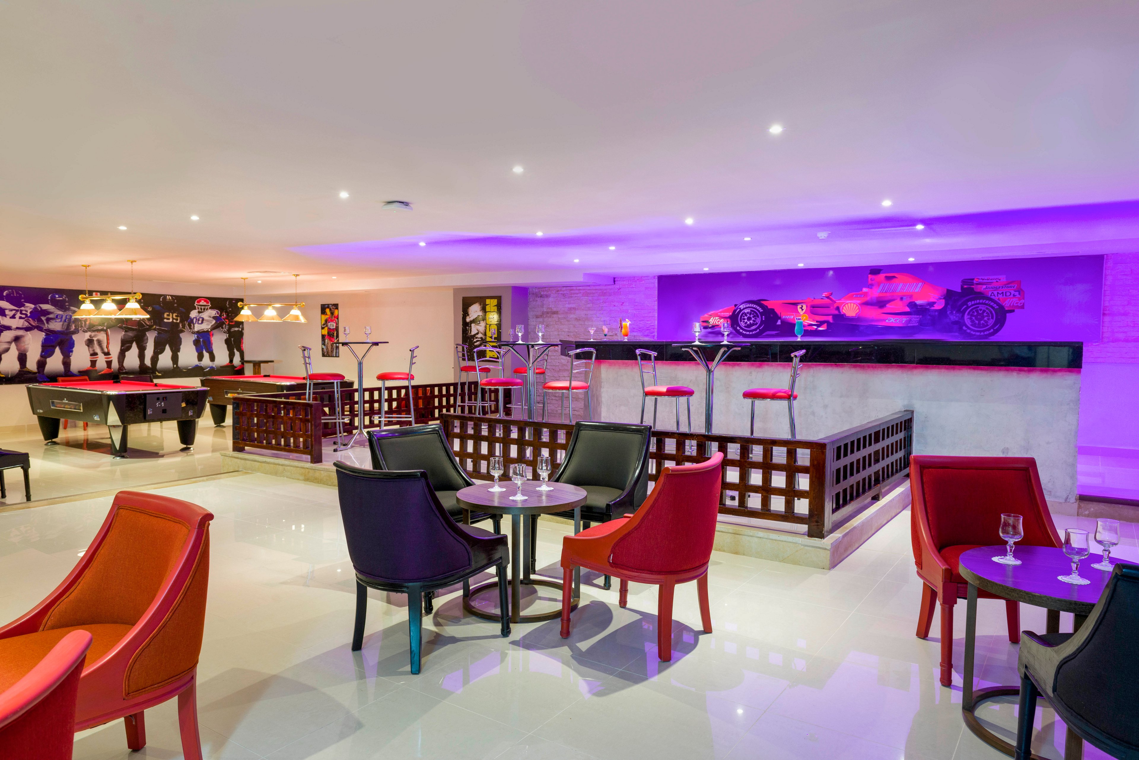 A contemporary lounge area features colorful armchairs and small round tables with wine glasses arranged on a glossy floor. In the background, several pool tables sit beneath warm pendant lights, creating a casual entertainment space. A raised bar area with high stools is highlighted by purple accent lighting. Large wall art of race cars and drivers reinforces the energetic, sports-themed atmosphere.