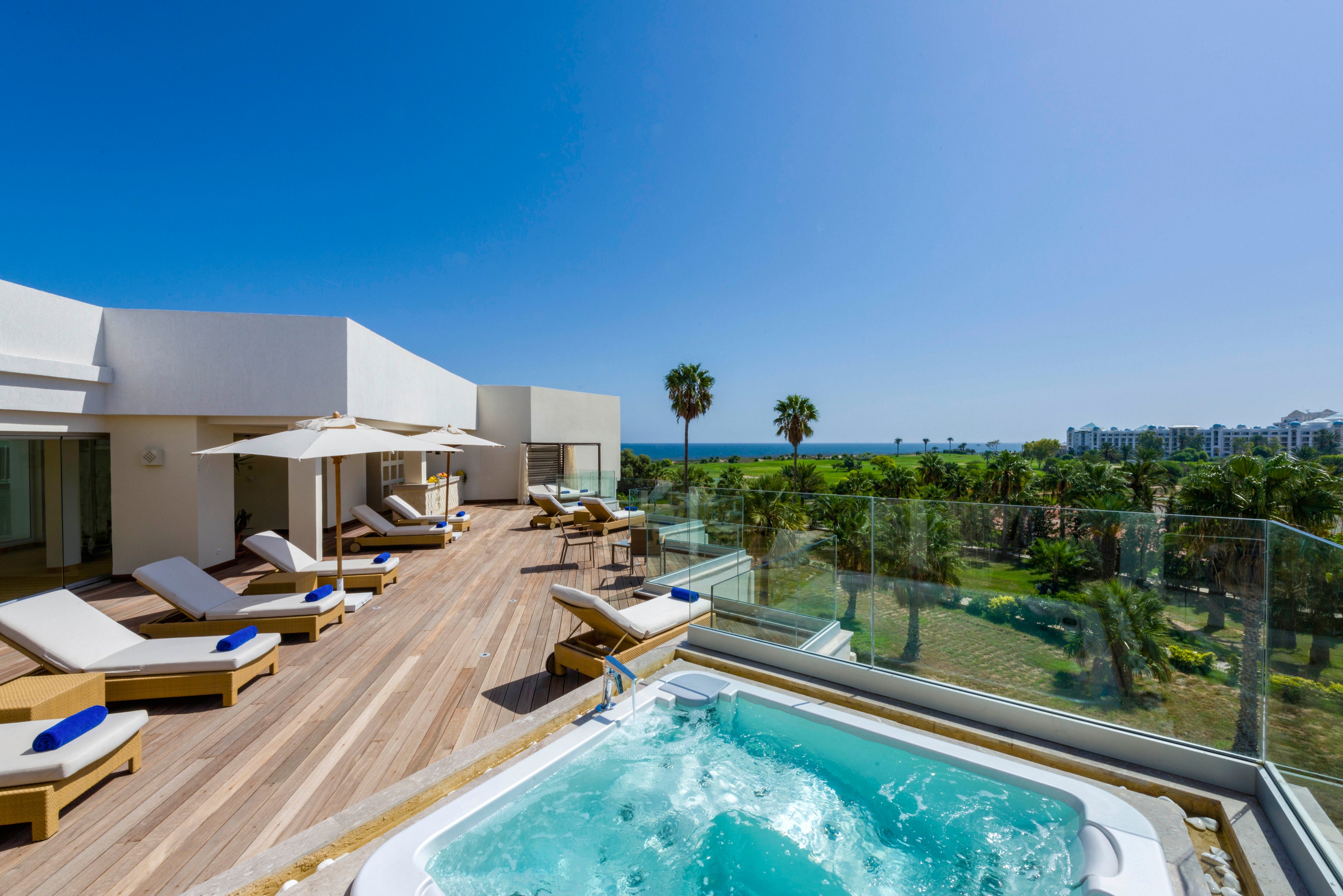 A modern rooftop terrace features a bubbling jacuzzi overlooking a lush coastal landscape. Wooden decking is lined with white cushioned sun loungers and large umbrellas, creating an inviting relaxation area. Glass railings frame the view of palm trees, green lawns, and distant resort buildings under a clear blue sky. The bright sunlight and clean architectural lines emphasize a serene, upscale vacation atmosphere.