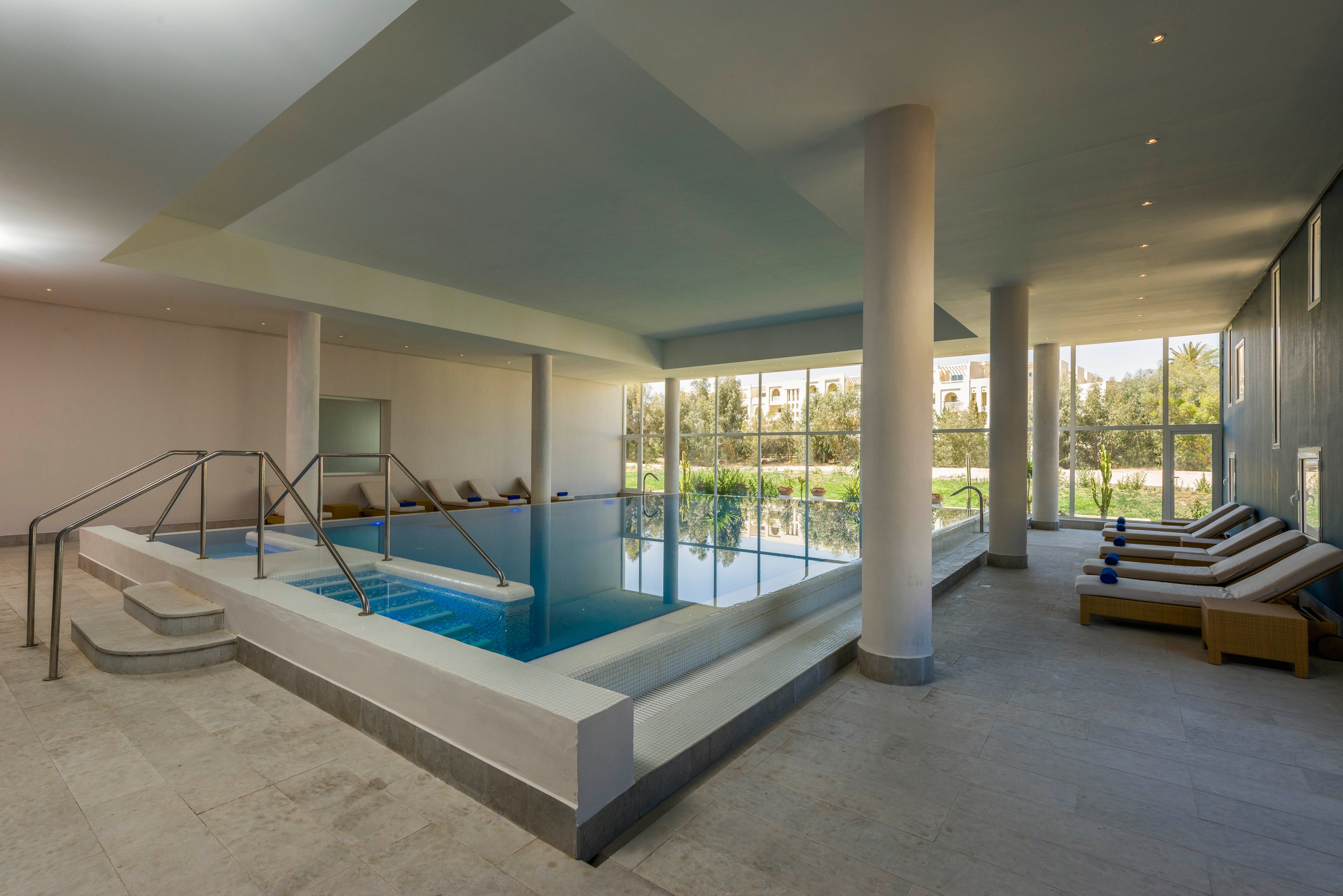 A modern indoor swimming pool is set within a spacious, minimalist room. Large floor-to-ceiling windows reveal greenery and buildings outside, filling the space with natural light. Several cushioned lounge chairs line the right side, while metal handrails provide access to the pool. The clean design and neutral colors create a calm, relaxing spa-like atmosphere.