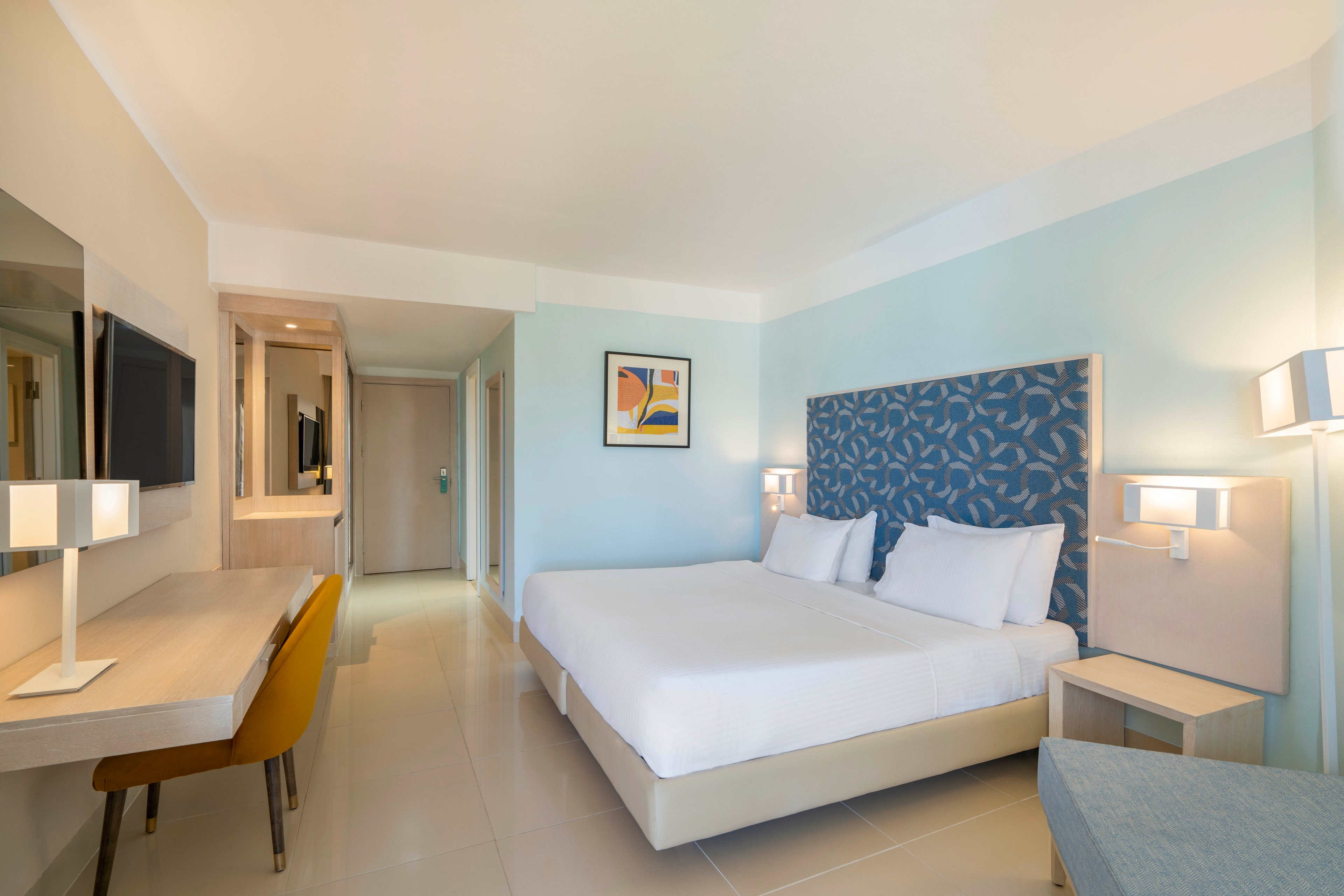 A contemporary hotel room features a large bed with a patterned blue headboard and crisp white linens. Soft blue and beige walls create a calm, coastal-inspired atmosphere. A sleek desk with a yellow chair faces a wall-mounted television, while matching bedside lamps provide warm lighting. The space appears clean, minimal, and inviting for travelers.