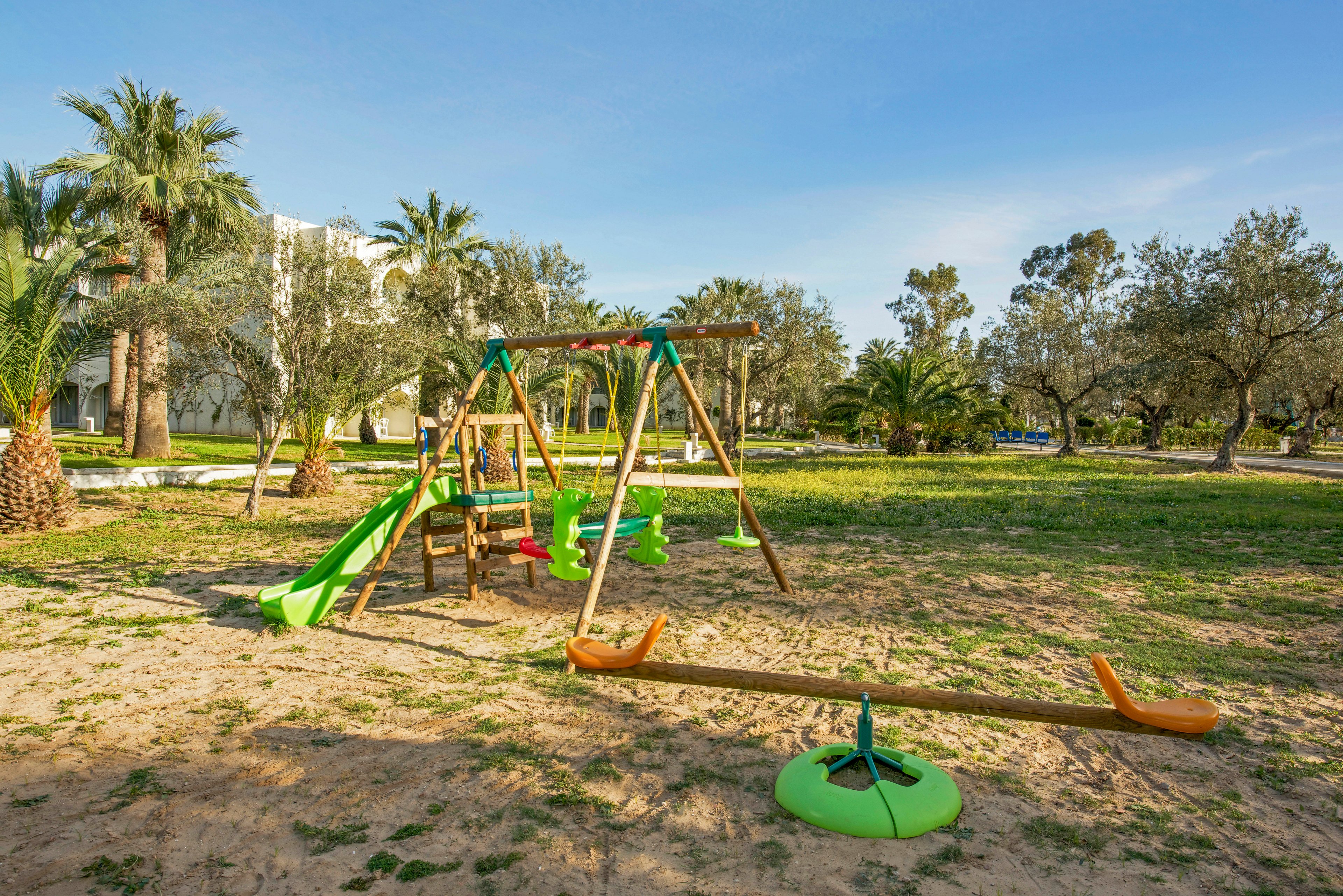 A wooden playground structure with slides and swings stands on a sandy area in an open park. The equipment includes bright green and orange plastic seats and a small round spinner. Surrounding the playground are grassy patches, palm trees, and other leafy trees under a clear blue sky. No children or adults are present, giving the scene a quiet, peaceful atmosphere.